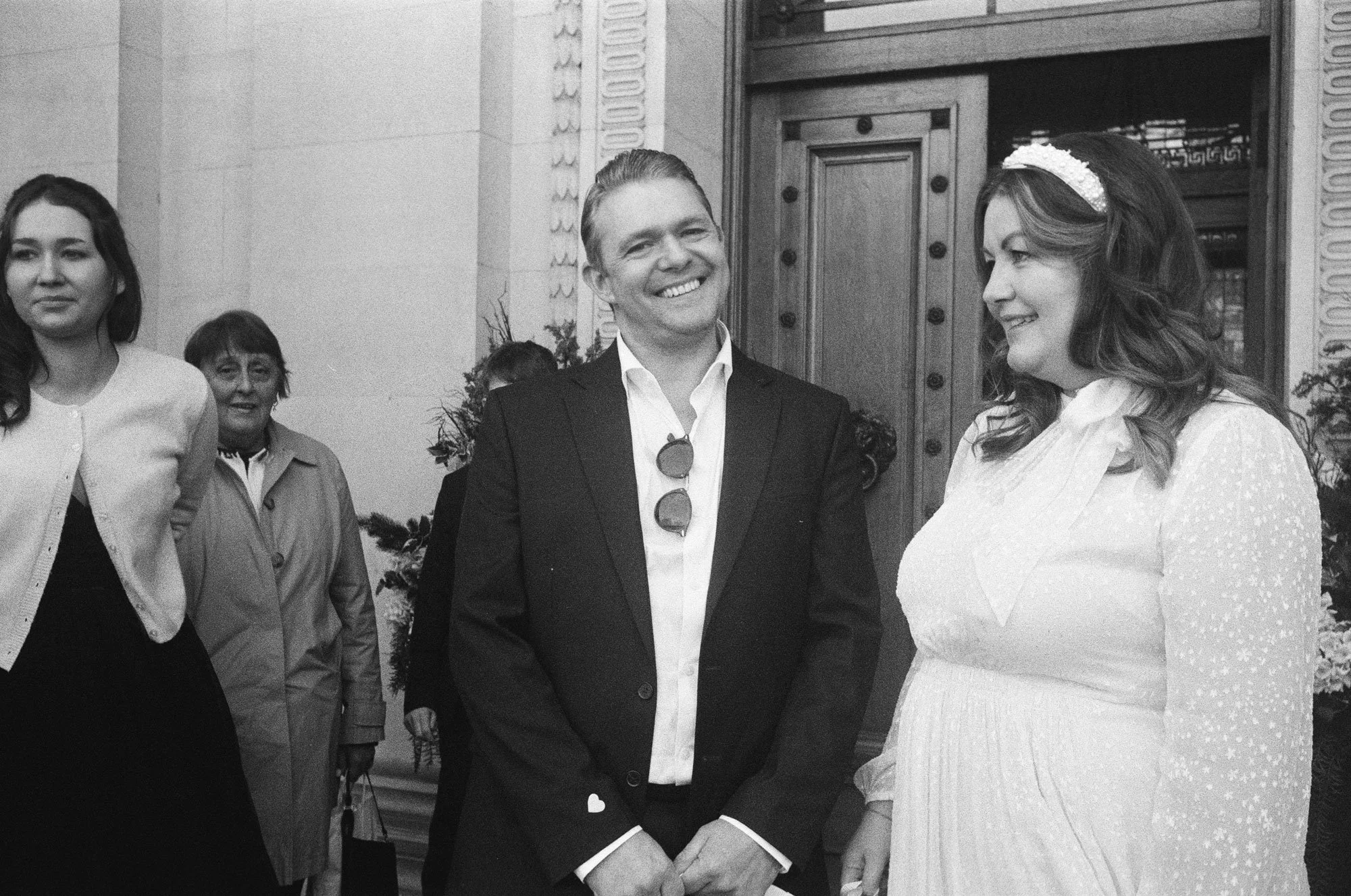 A wedding scene with a man in a suit and a woman in a white dress smiling at each other, surrounded by guests indoors.