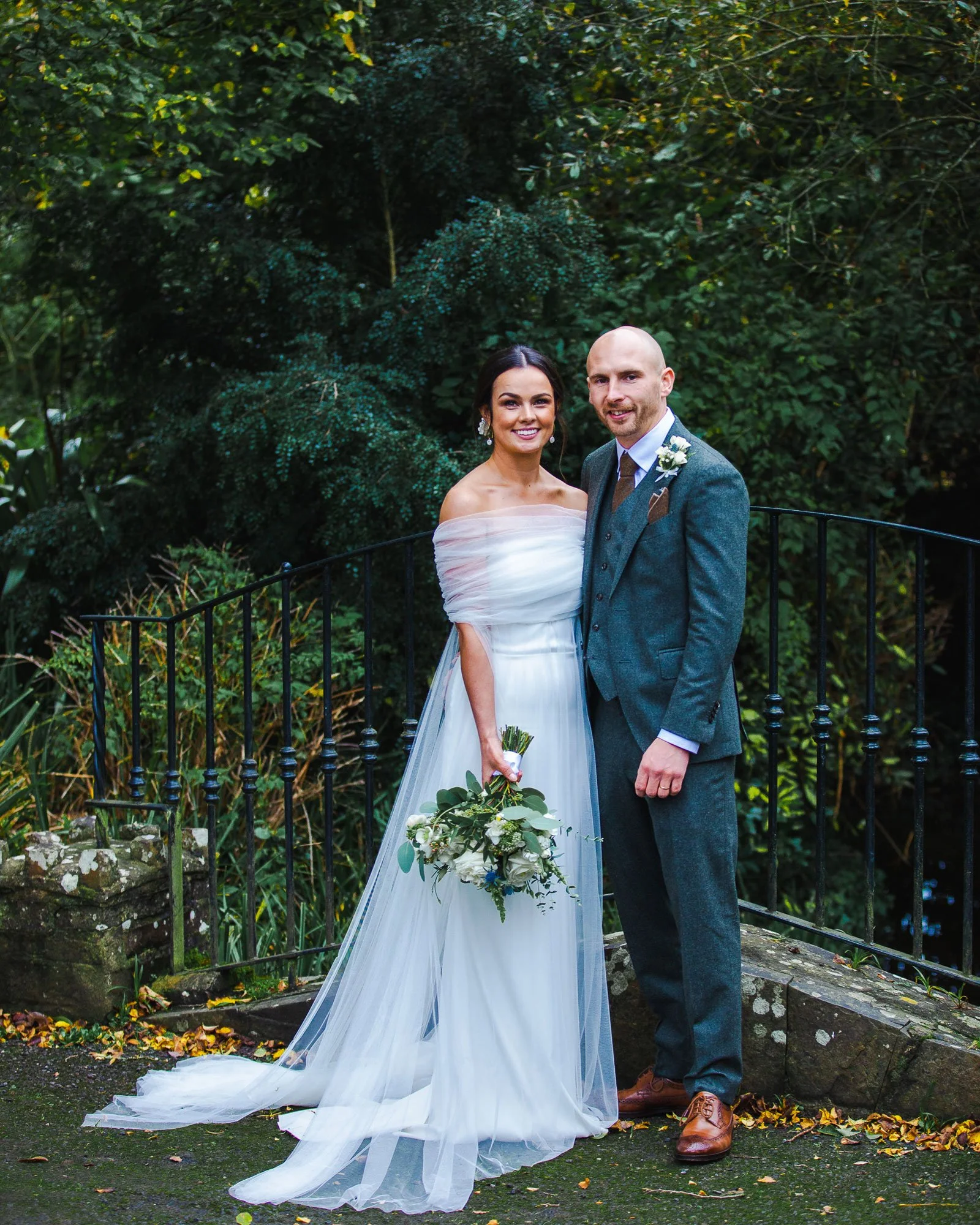 A bride and groom pose for a wedding photo outdoors, with lush green foliage in the background. The bride wears a white off-shoulder gown and holds a bouquet, while the groom wears a gray suit with a boutonniere.