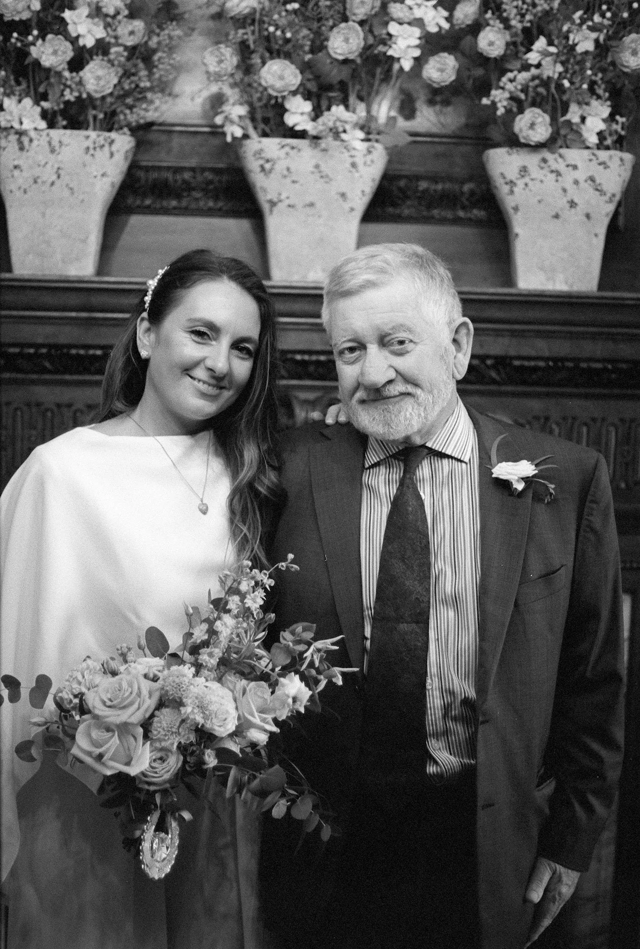 A young woman in a white dress holding a bouquet, standing next to an older man in a suit with a boutonnière, both smiling in an indoor setting with flower arrangements behind them.