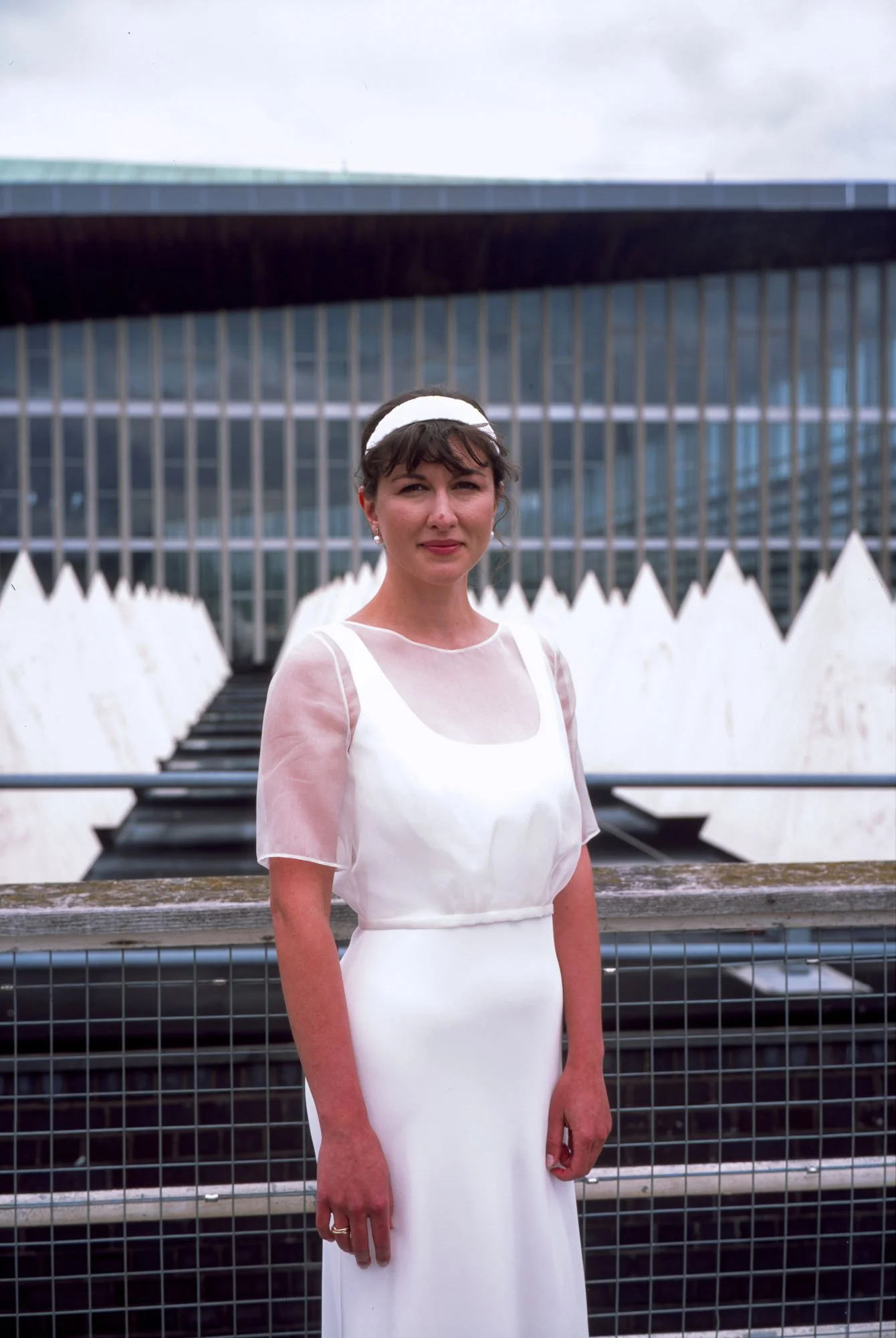 A woman in a white dress standing outdoors in front of modern glass building and white water fountains.