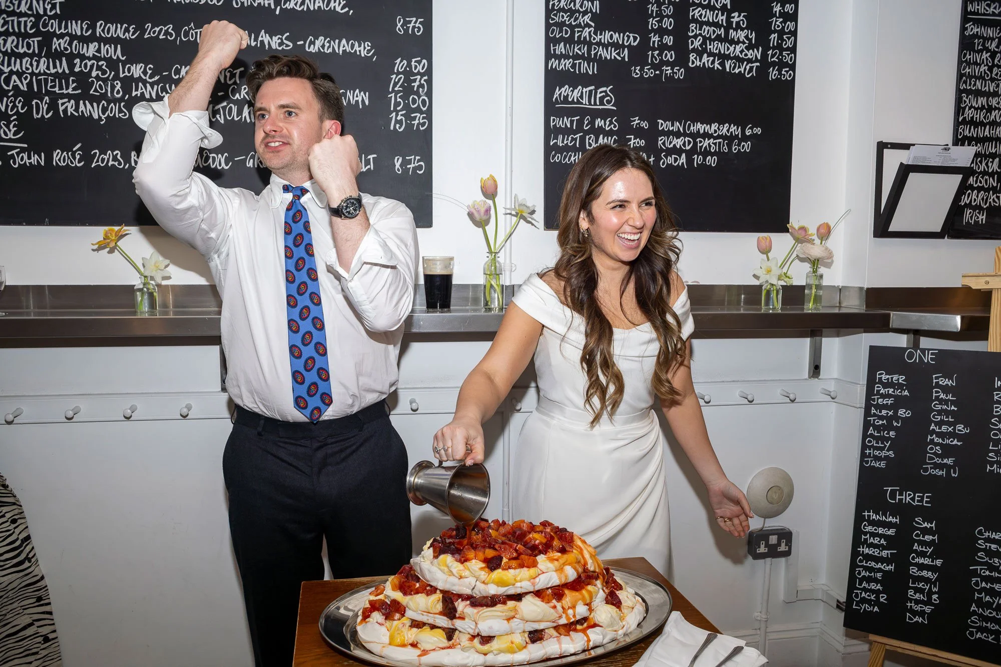 A man and woman celebrating at a gathering, with the woman pouring syrup on a large meringue dessert topped with fruit, and the man adjusting his tie.