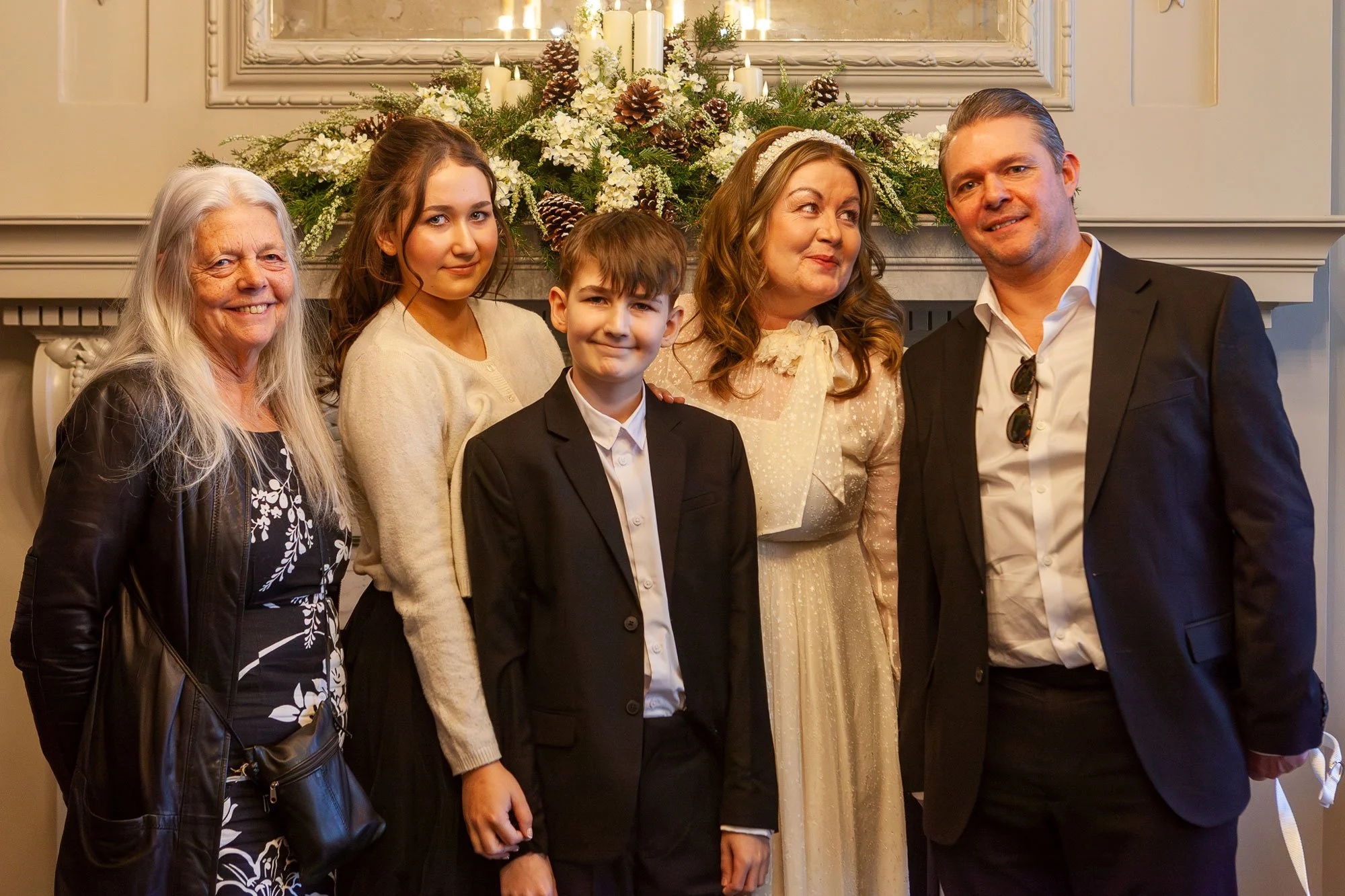 Family posing together at a holiday gathering in front of a decorated fireplace mantel with white flowers, pinecones, candles, and Christmas ornaments. The group includes an elderly woman, two women, two children, and two men, all dressed in festive 