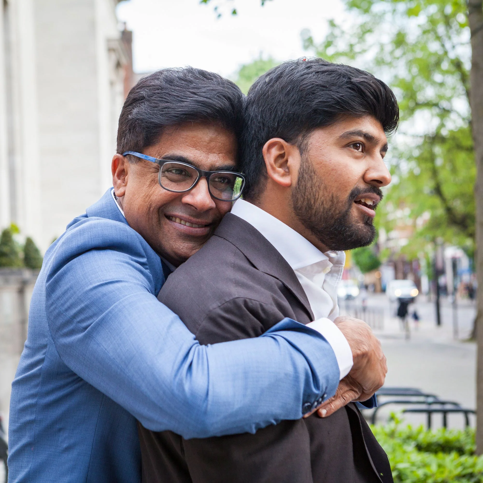 A man in a blue suit hugging another man in a dark suit on a city street with trees and buildings in the background.