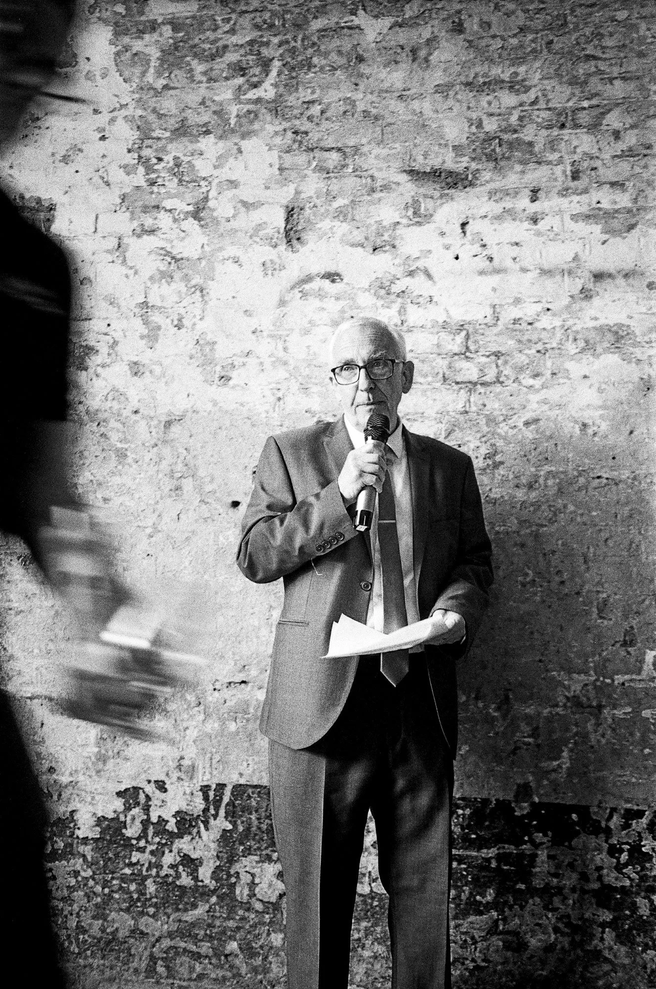 A black and white photo of an older man in a suit and tie speaking into a microphone while holding papers, standing against a textured brick wall.