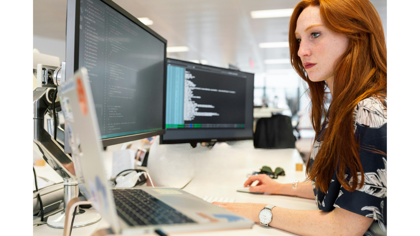 A busy woman with red hair and a floral shirt working on a computer with two monitors displaying coding or data.