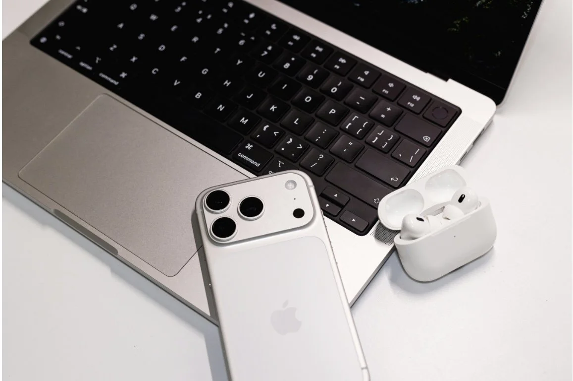 A silver MacBook with black keyboard, white iPhone with three camera lenses, and white Apple AirPods in their charging case on a white surface.