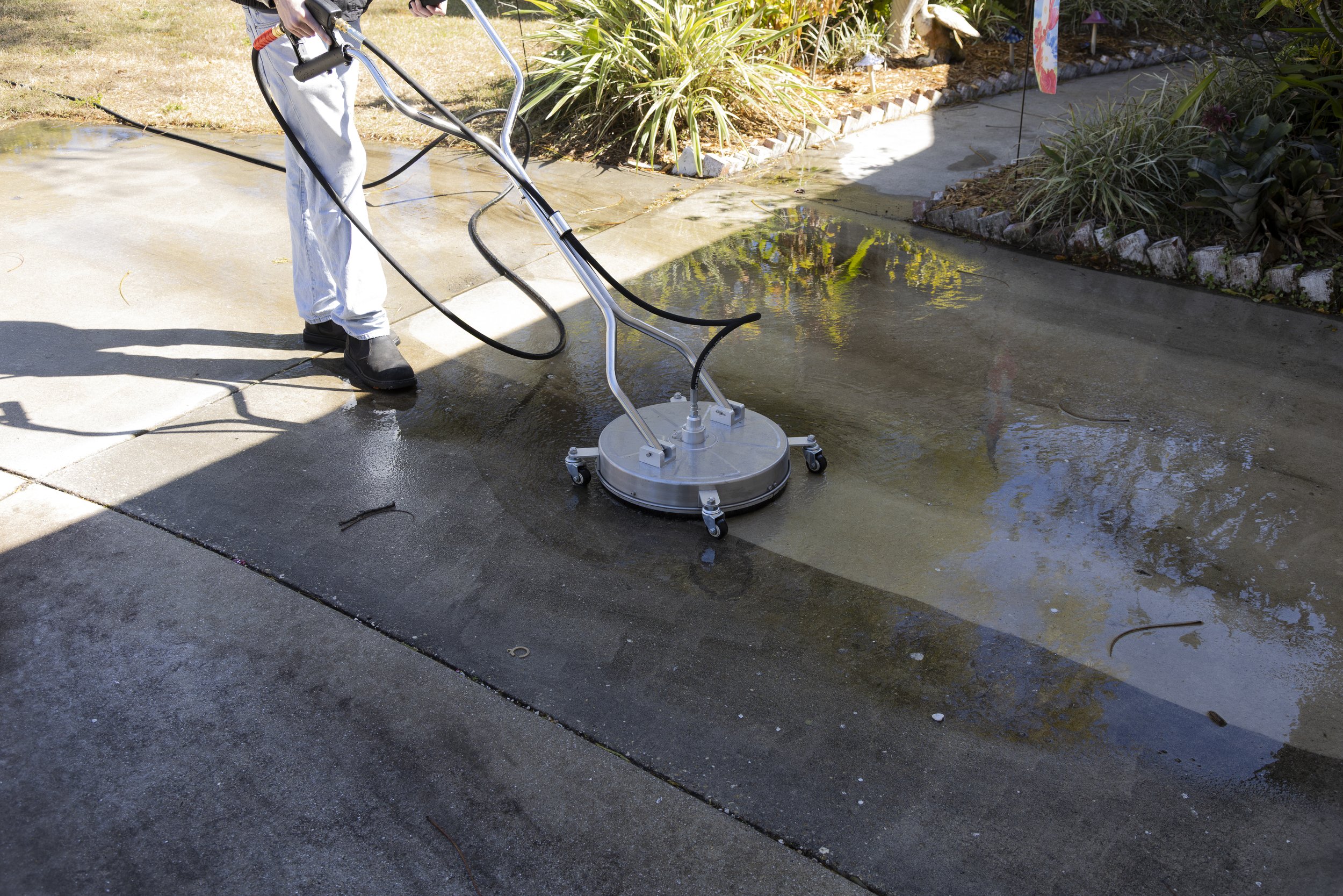 Person using a floor scrubber machine to clean concrete driveway outside on sunny day.