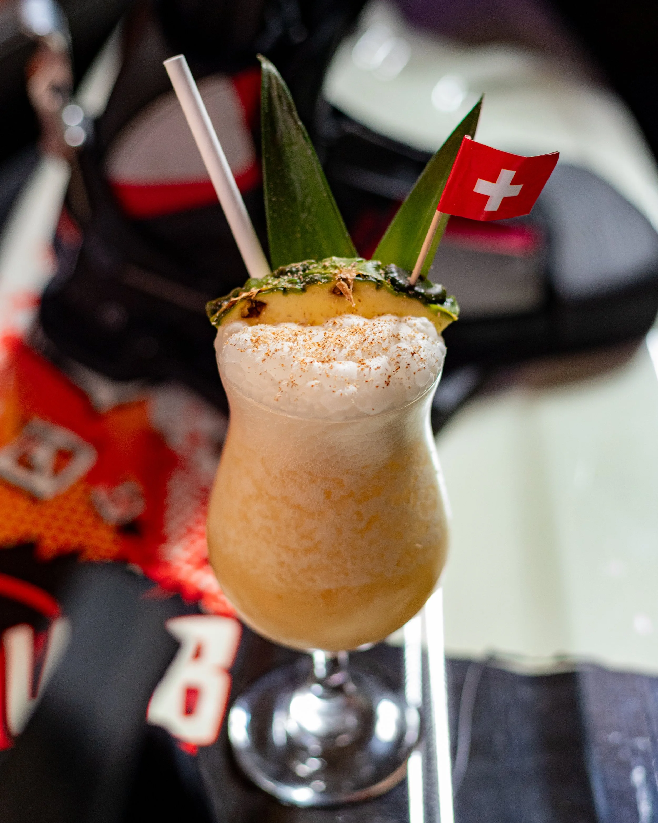 A tropical cocktail in a tall glass decorated with a pineapple slice, two green leaves, and a Swiss flag toothpick. The drink is creamy and sprinkled with cinnamon or nutmeg on top.