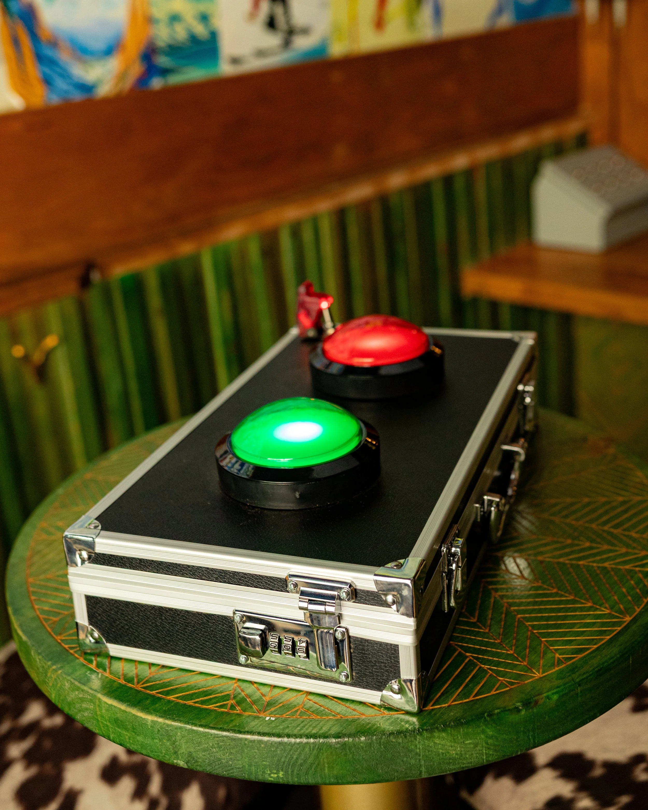 A black carrying case with metal accents on a green table. On top of the case, there are two large colored buttons, one red and one green, with the green button illuminated.