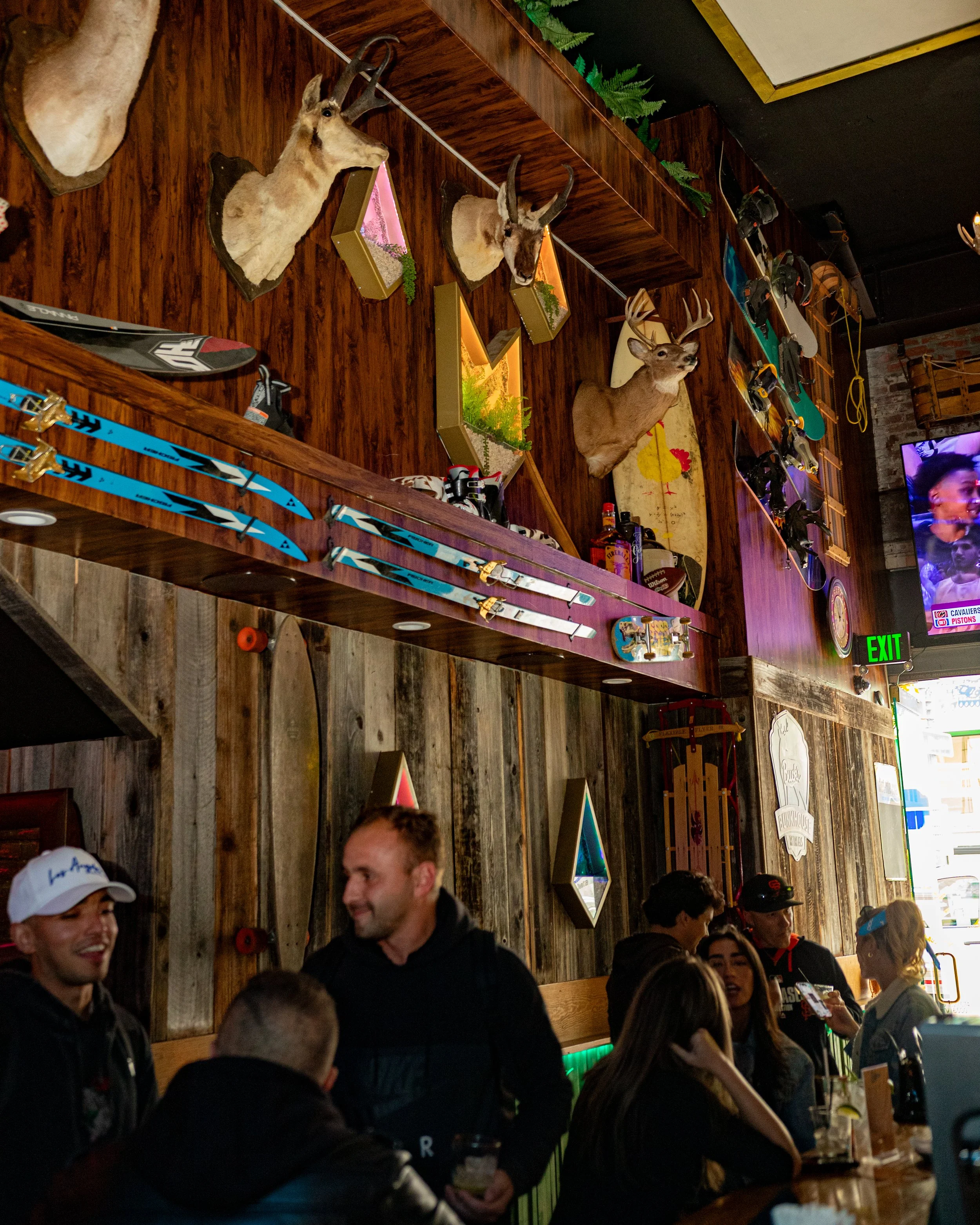 A lively bar or restaurant with a wooden interior decorated with mounted animal heads including deer and antelope, and American flags. People are gathered around the bar, chatting and enjoying drinks. Ski equipment such as skis is displayed along the