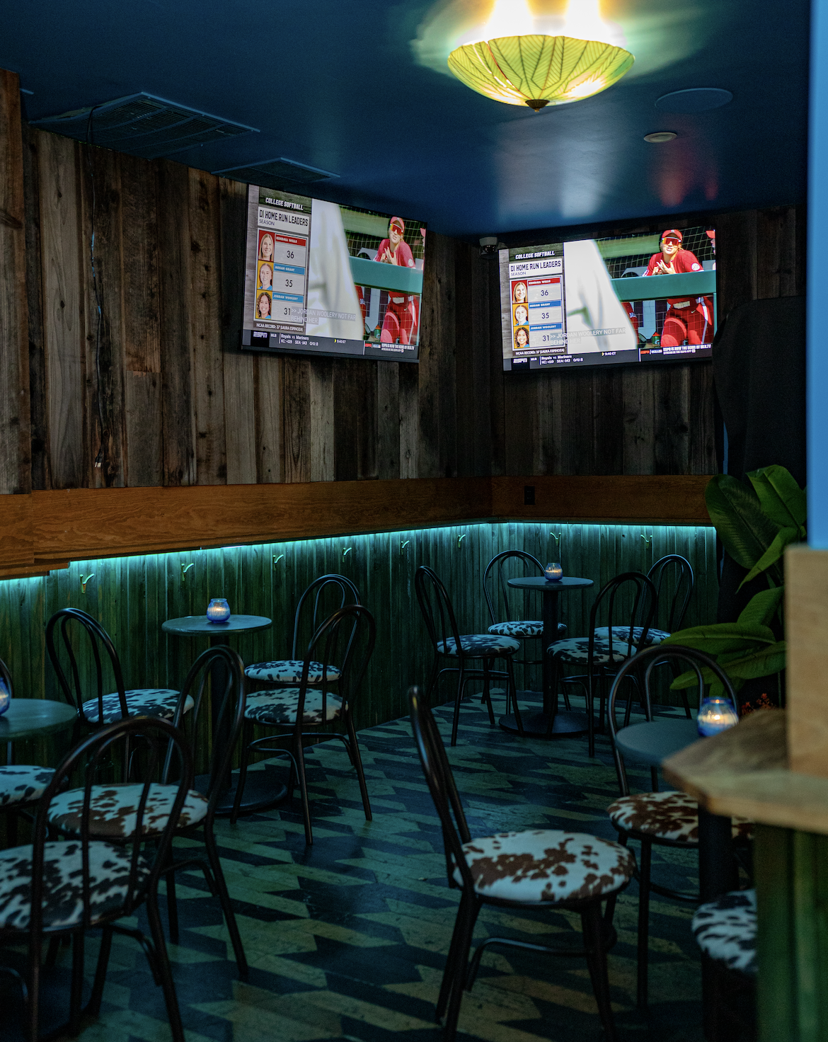 Empty bar or lounge area with black chairs with patterned cushions around small round tables, lit by candles. Two television screens mounted on a wooden wall display sports scores and athletes. The ceiling has a yellow-green lampshade, and there is g