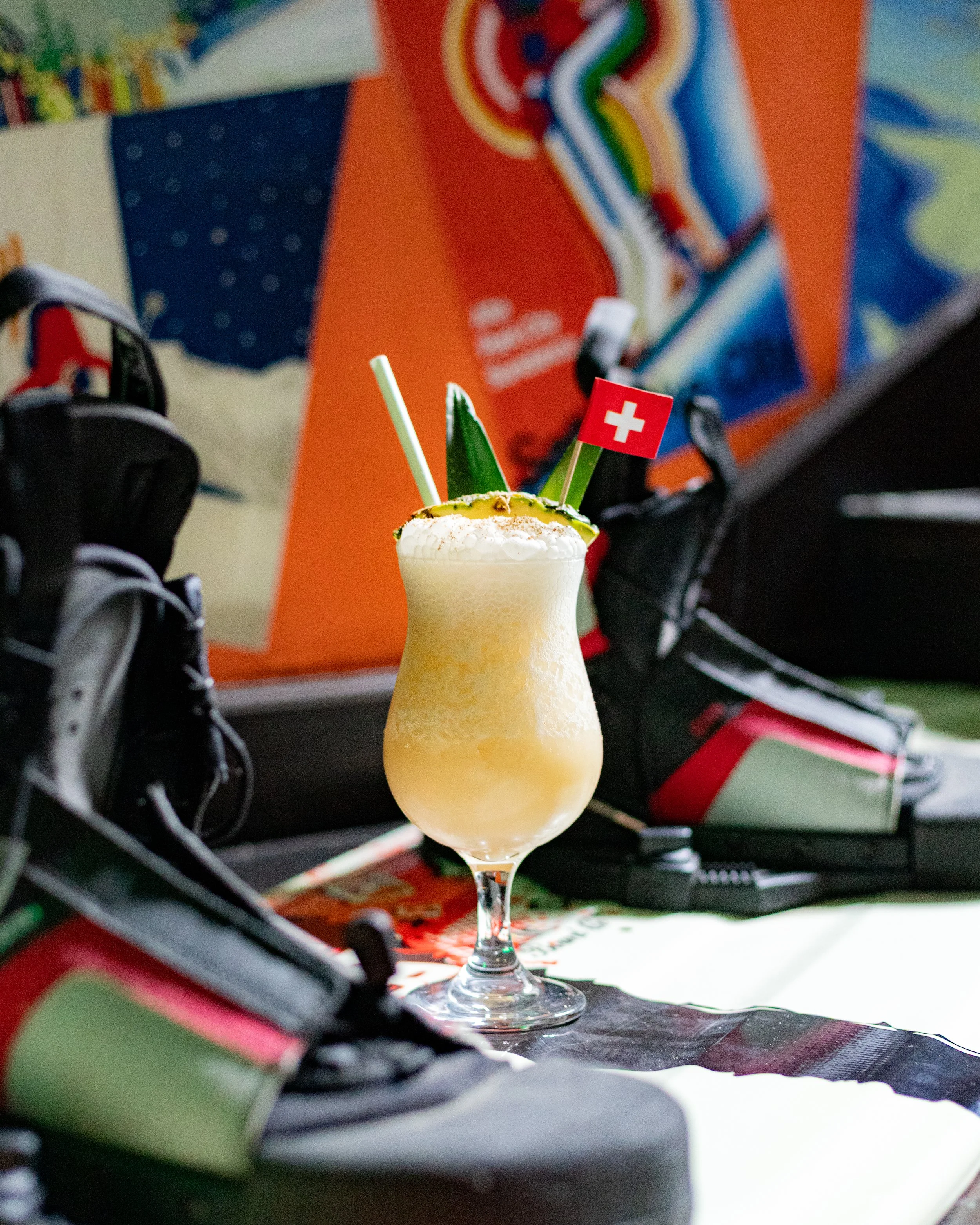 A colorful cocktail in a tall glass with a long straw, decorated with small flags of Switzerland and Italy, placed on a cluttered desk with athletic shoes and colorful posters in the background.