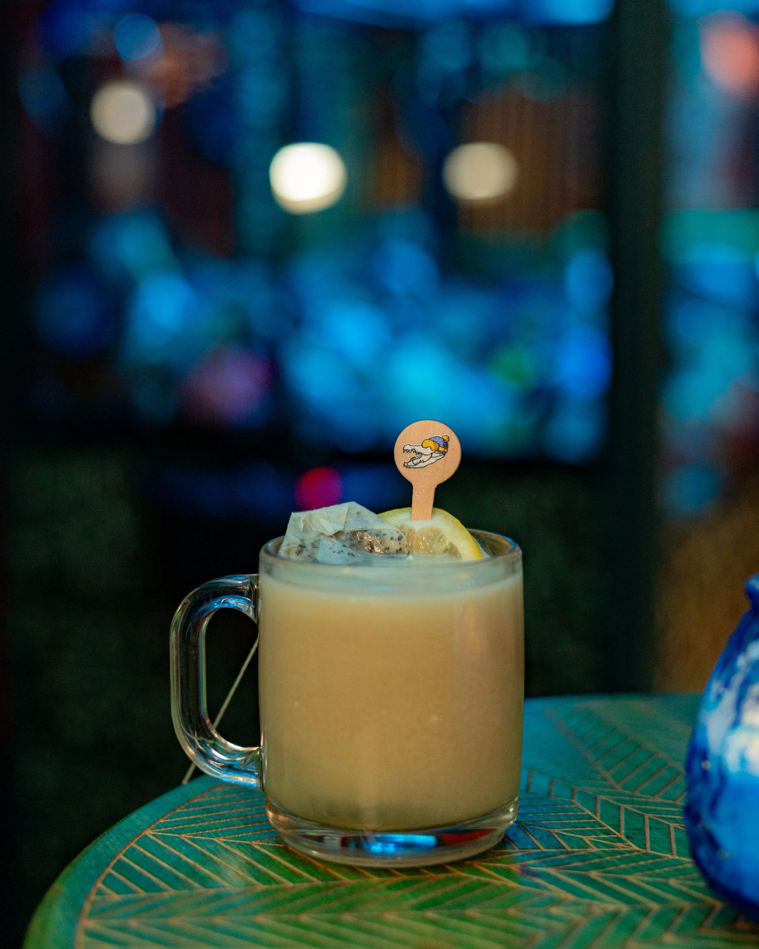 Close-up of a glass mug with a hot beverage garnished with a lemon wedge and a paper umbrella in a dimly lit setting with defocused colorful lights in the background, placed on a decorative teal plate.