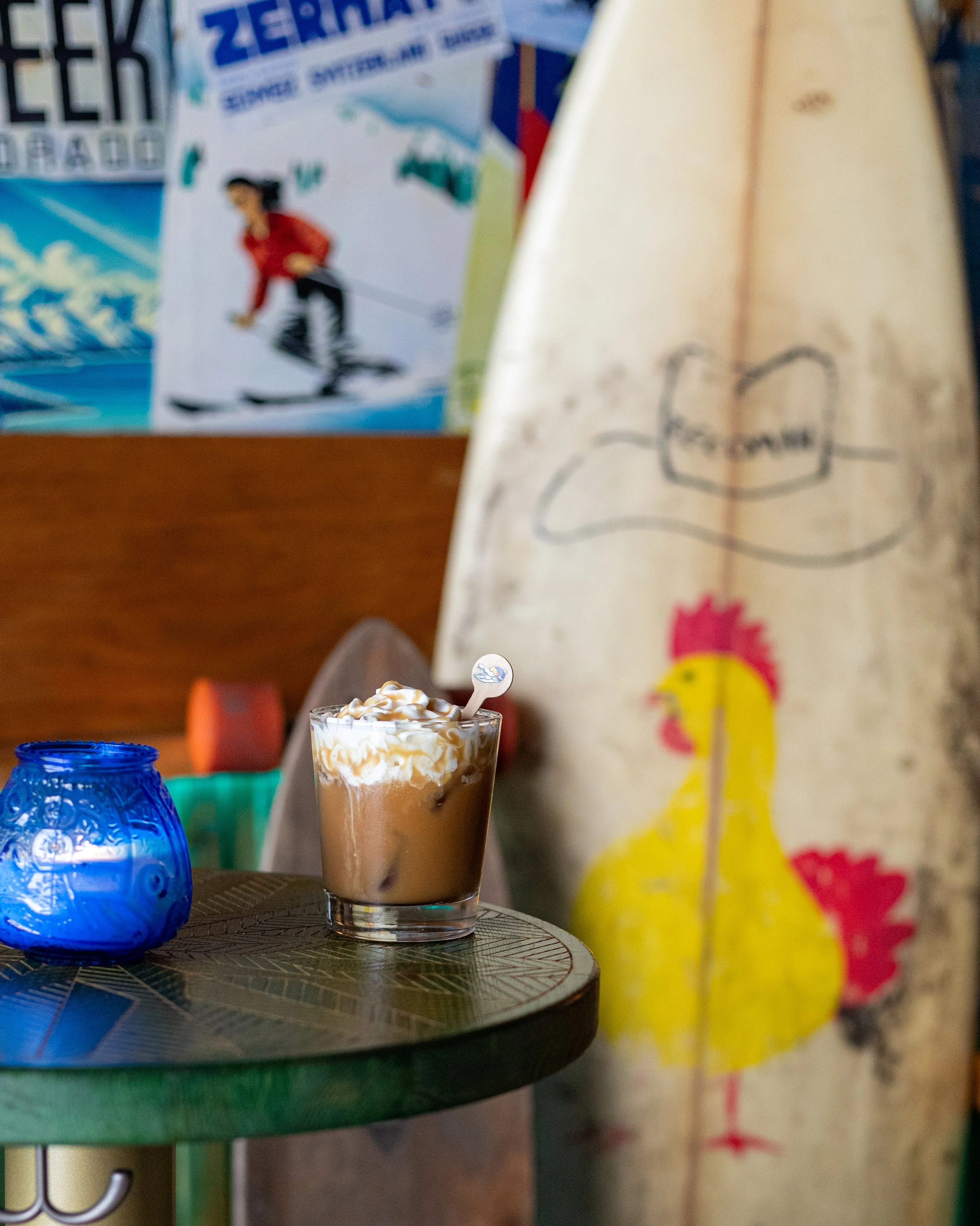 A glass of iced coffee with whipped cream on top sits on a table. There is a blue decorative jar and a paper bag with a colorful design in the background.