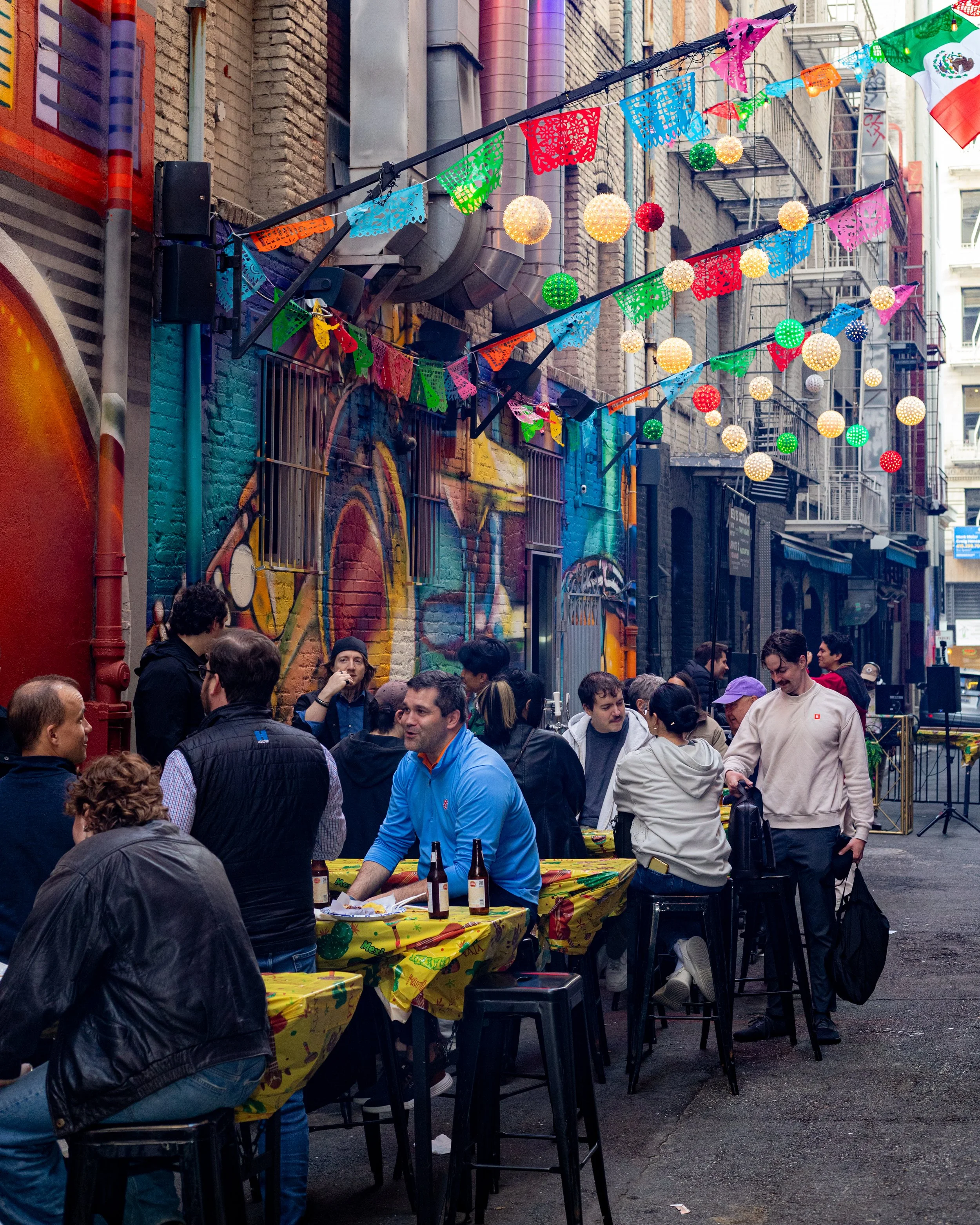 A street scene with people gathered around tables covered with yellow tablecloths decorated for a celebration, with colorful festive banners and lights hanging overhead. The background features brick buildings, some with murals and fire escapes.