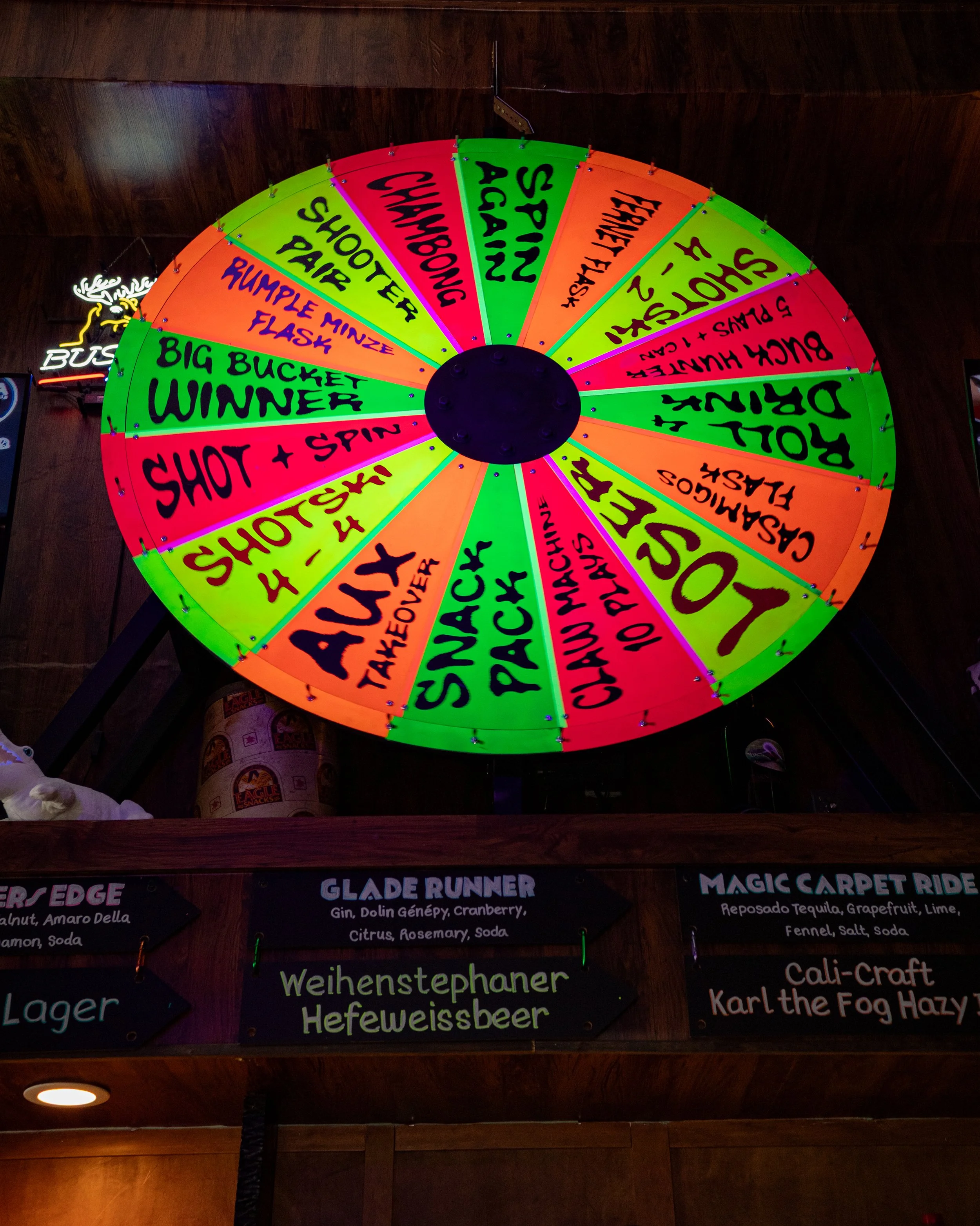 Colorful spinning wheel with various game or drink options written on each segment, attached to a wooden ceiling.