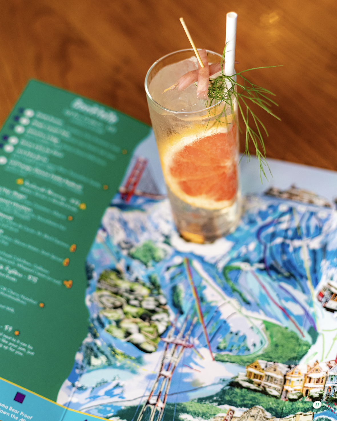 A tall glass of grapefruit soda garnished with a slice of grapefruit, a sprig of dill, and two straws, placed on a table with a colorful map and a green menu.