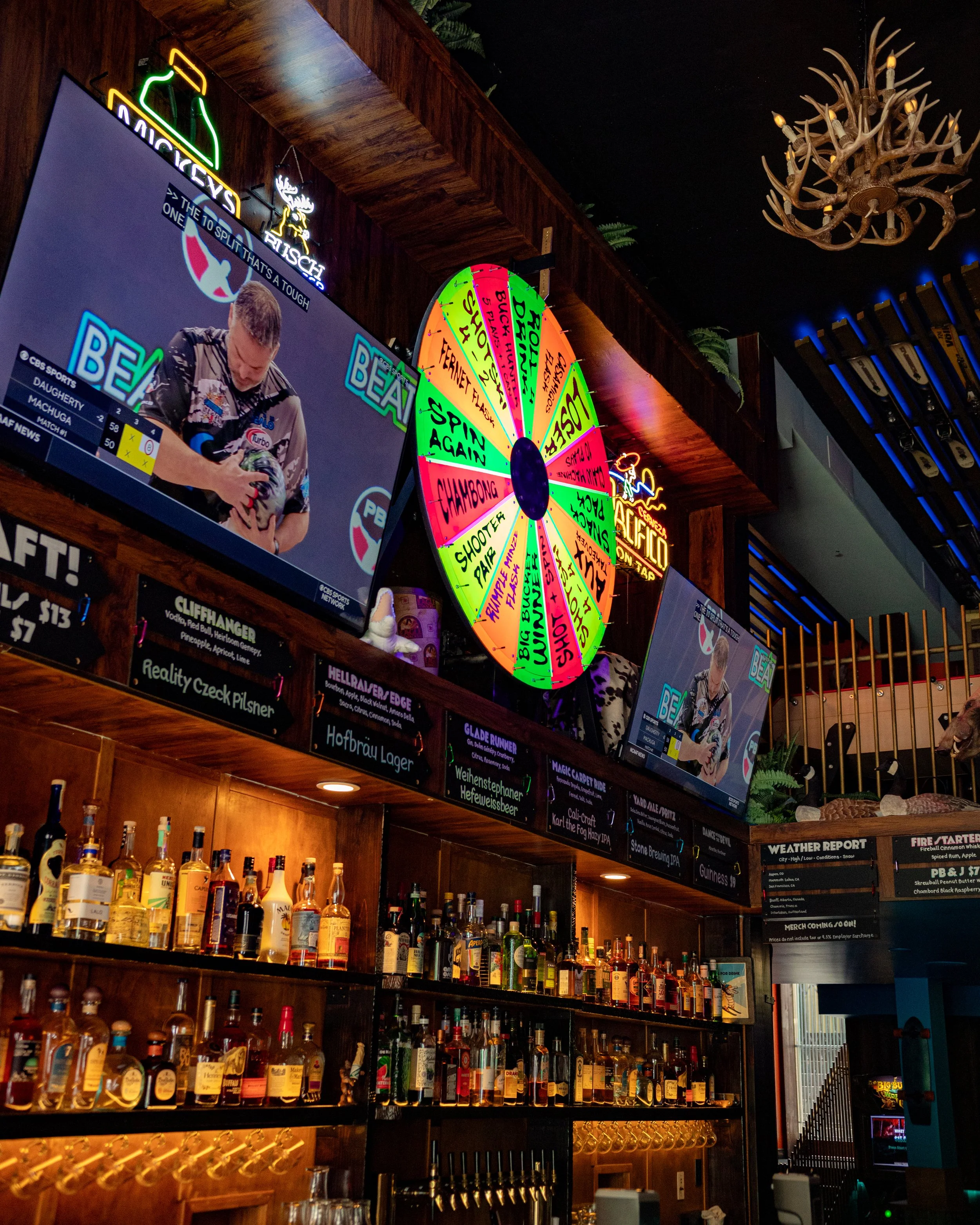 A bar interior with a neon sign, a digital display, a colorful spin wheel, bottles of alcohol on shelves, and a chandelier made of antlers.