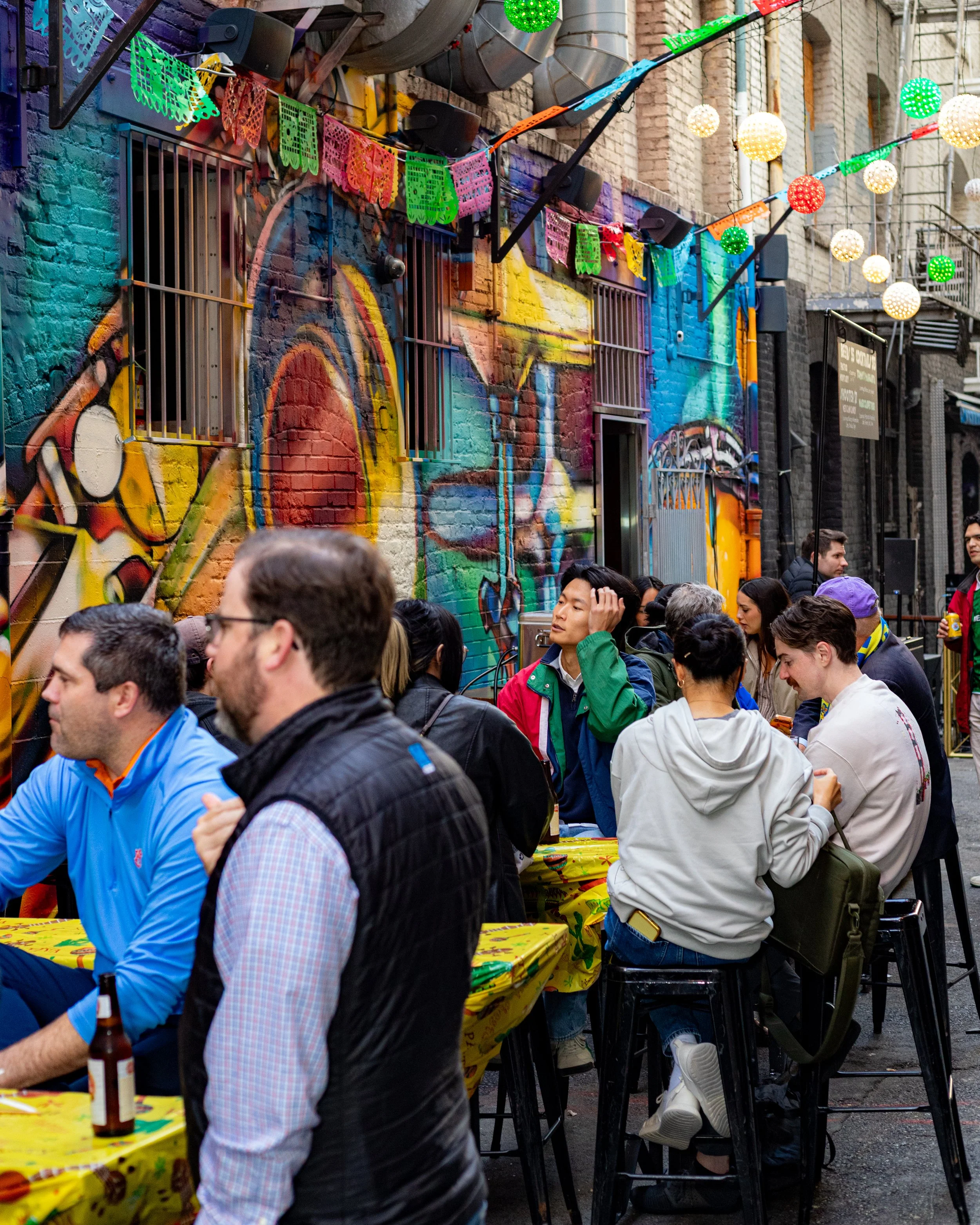 People gathered at an outdoor celebration with colorful papel picado banners and festive string lights on a brick building backdrop.