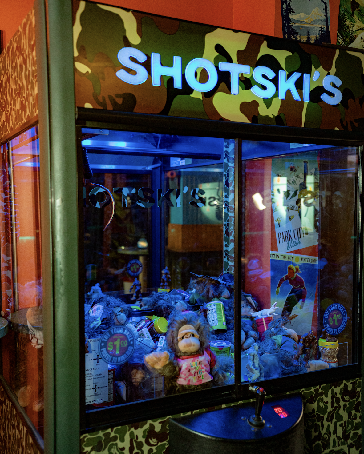 A claw machine labeled 'Shotski's' filled with plush toys and cans of Pringles, with a colorful camouflage design on the top edge and tropical-themed artwork on the walls.