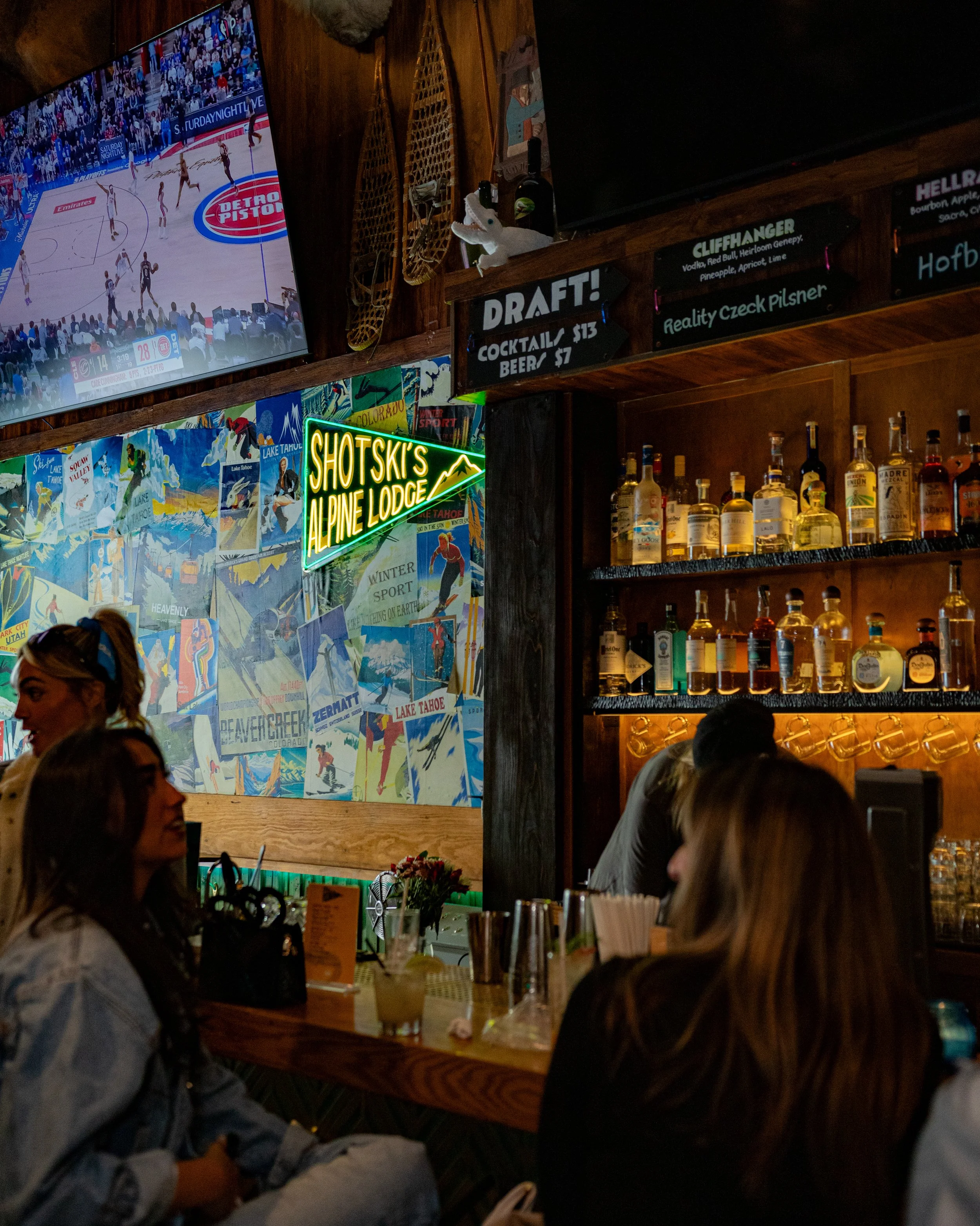 Bar scene with people sitting at the counter, various liquor bottles on shelves, illuminated neon sign reading "Shotski's Al Pine Lodge," and a television showing a basketball game.