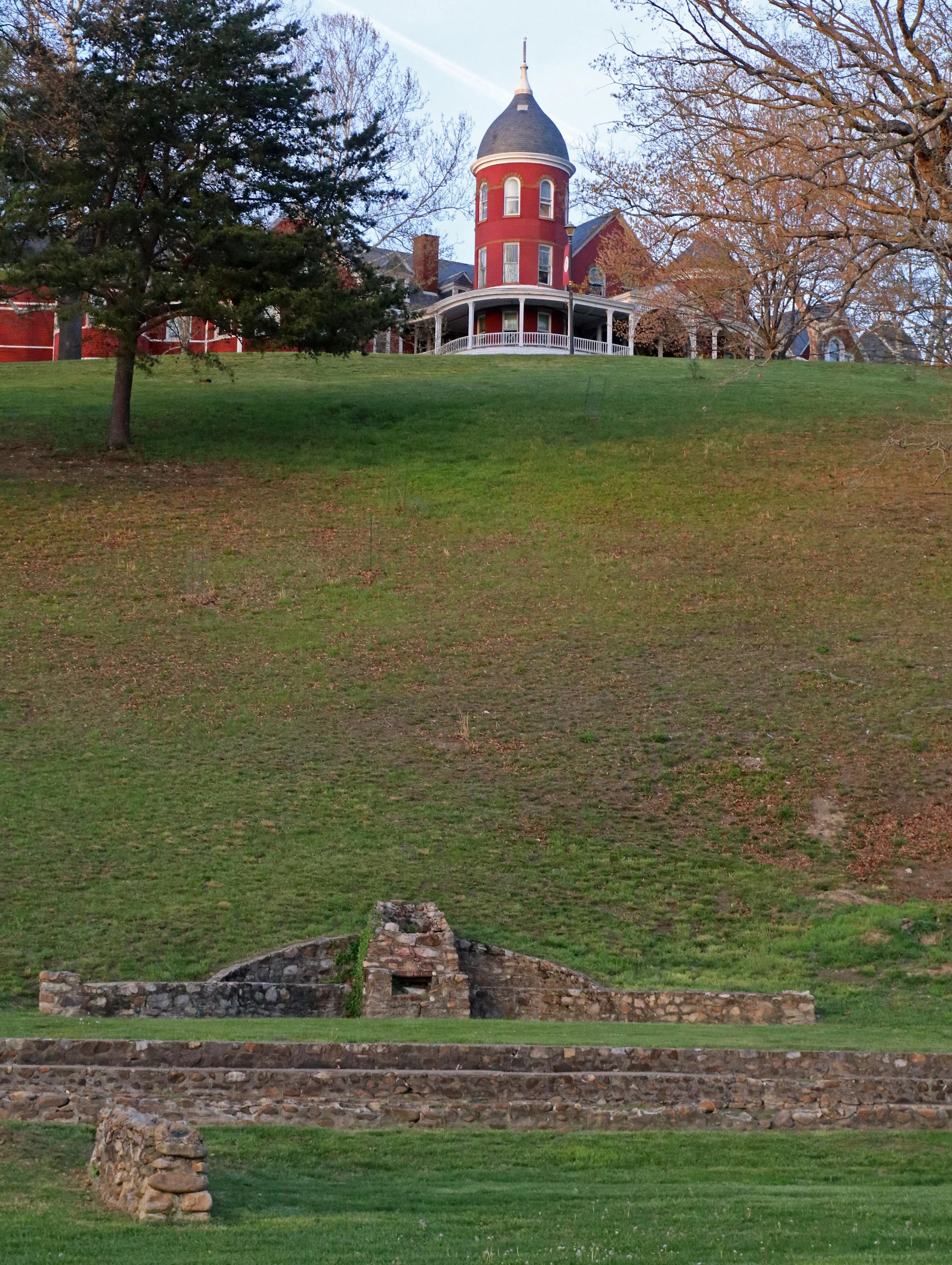Large red turrent from mansion sized building on top of a steep hill. Stone fence & structure at bottom of hill