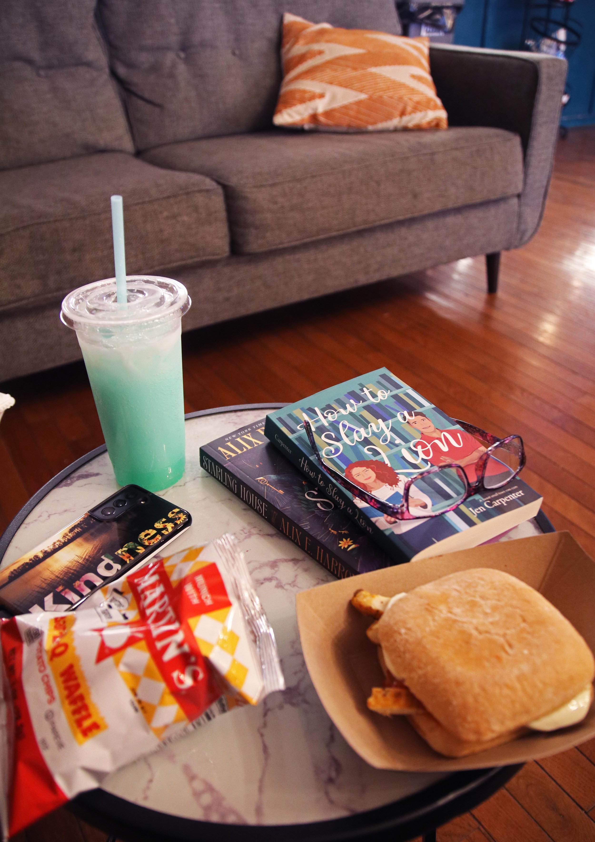 Small round table in Straws soda shop with Salty Dog drink, two books stacked with reading glasses on top, sandwich, chips and cell phone, couch in background
