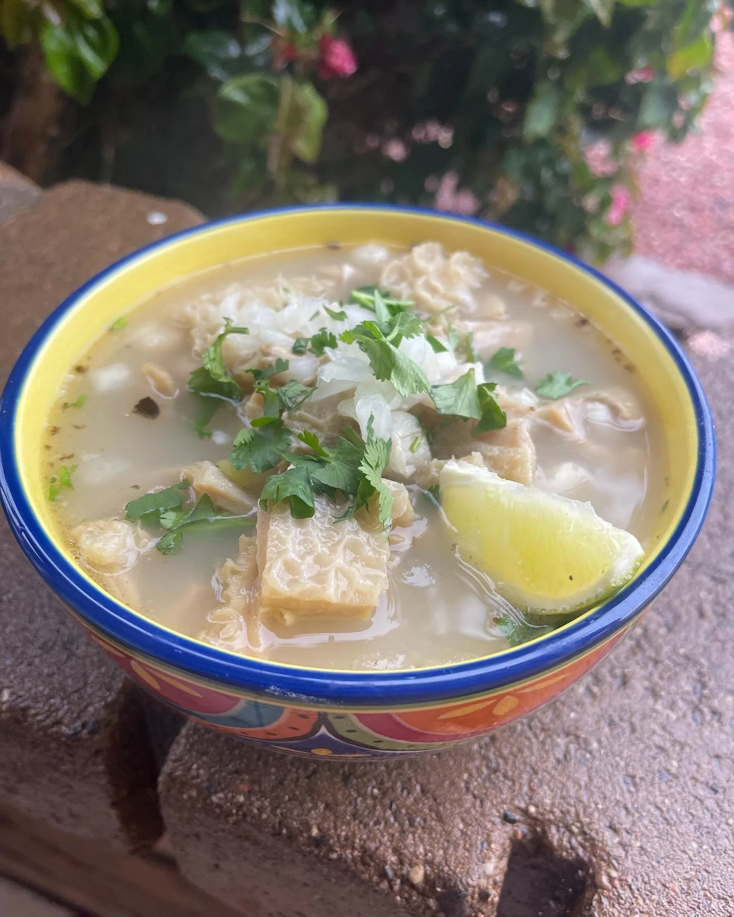 Bowl of fish soup garnished with cilantro and lime wedge, placed on a brick surface outside.