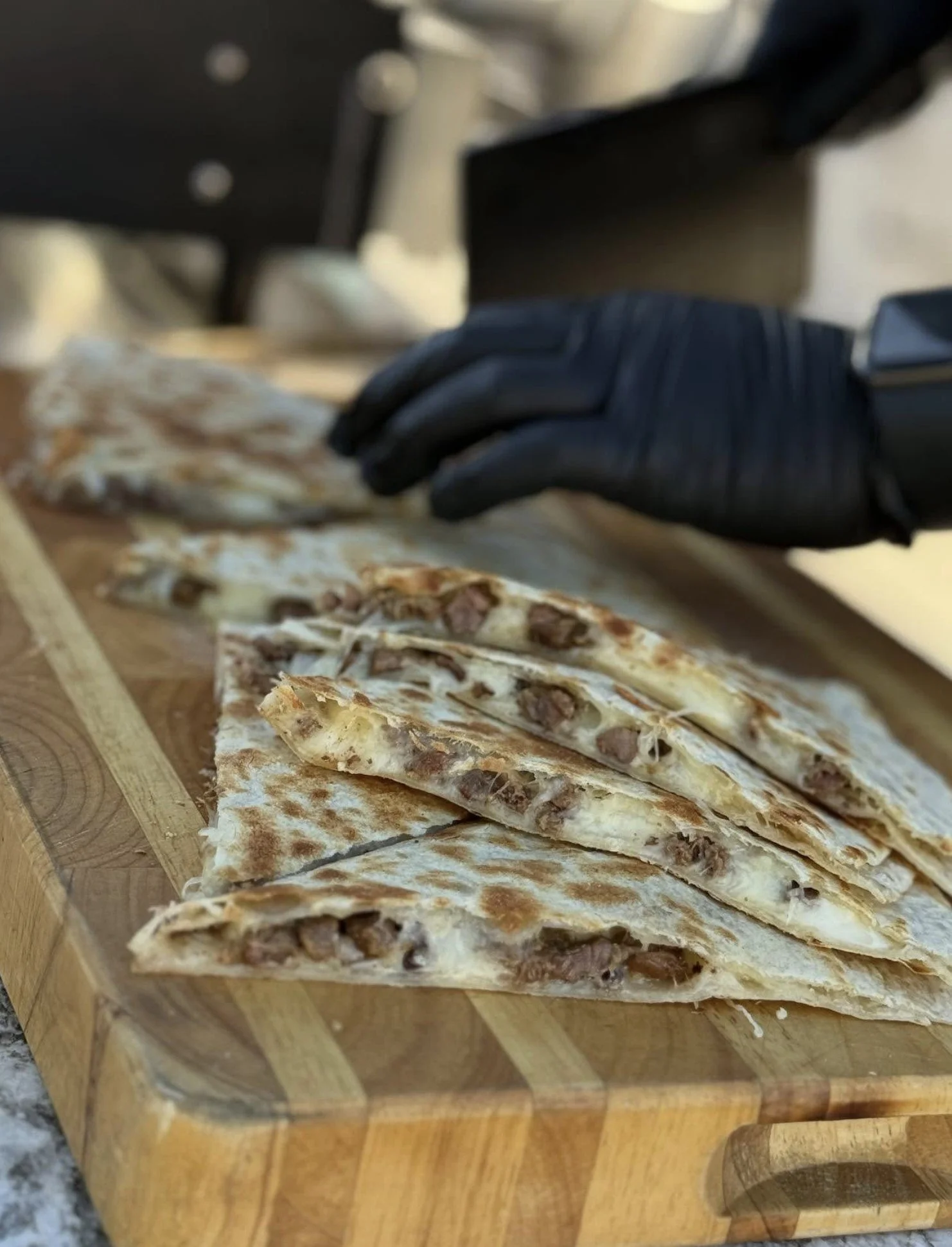 Sliced beef and cheese quesadillas on a wooden cutting board, a person is slicing them with a black gloved hand.