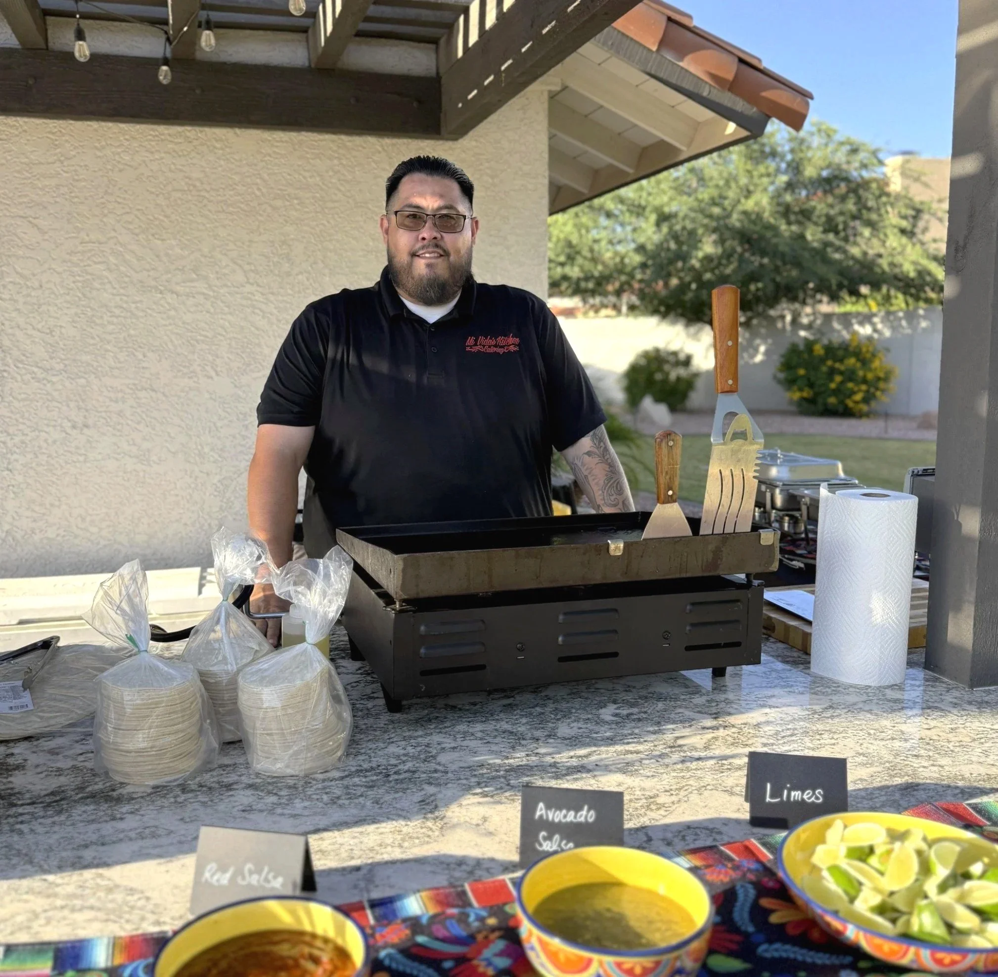 Man standing behind a table at an outdoor food stall or event, with a grill, tongs, spatula, and paper towels on the table. Buckets of tortilla wraps and bowls of salsa, avocado, and limes are visible in the foreground. Bright sunny day with greenery and house in the background.