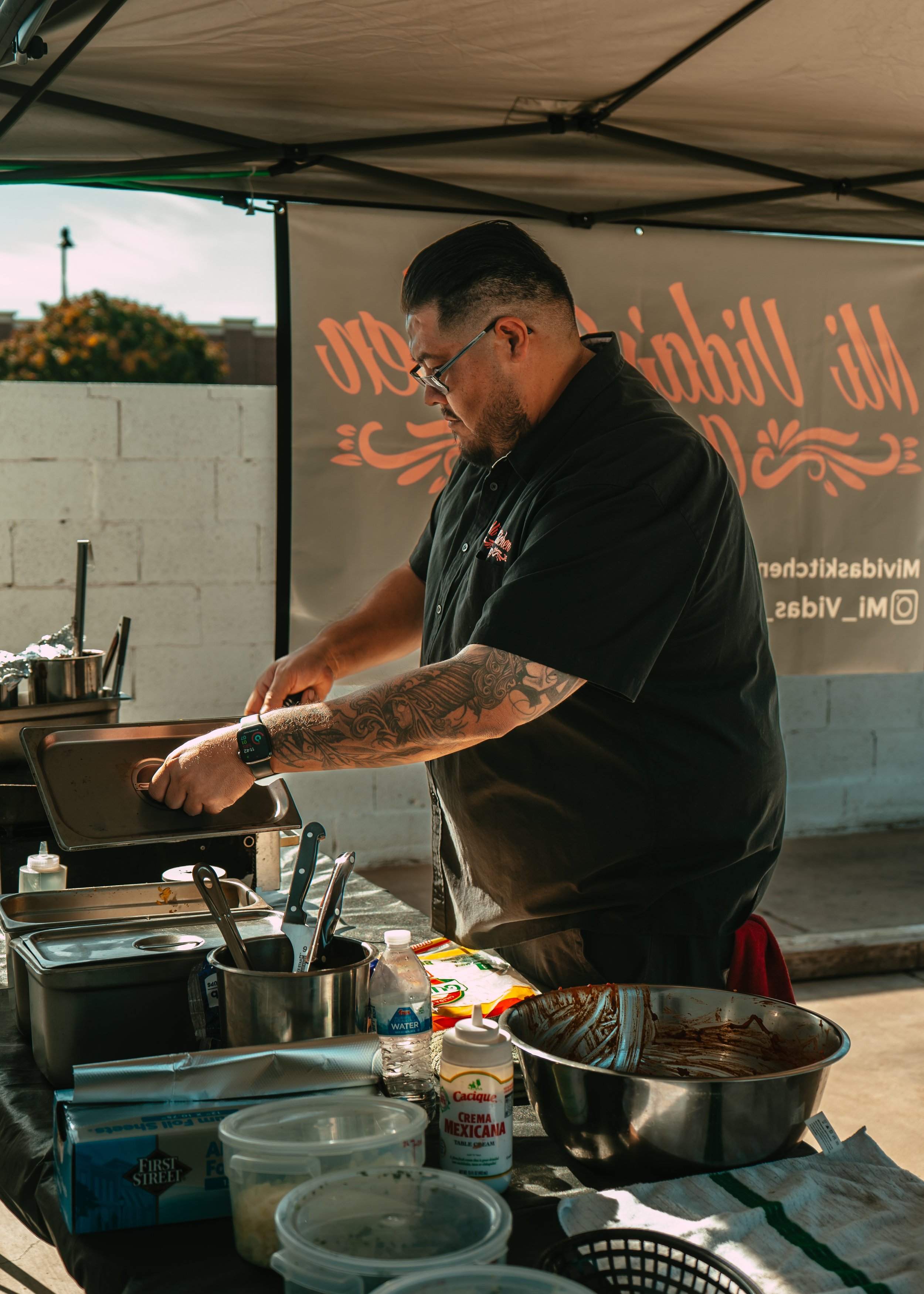 A man with tattoos and glasses wearing a black shirt working at an outdoor food stand, preparing food with various ingredients and utensils, under a canopy.