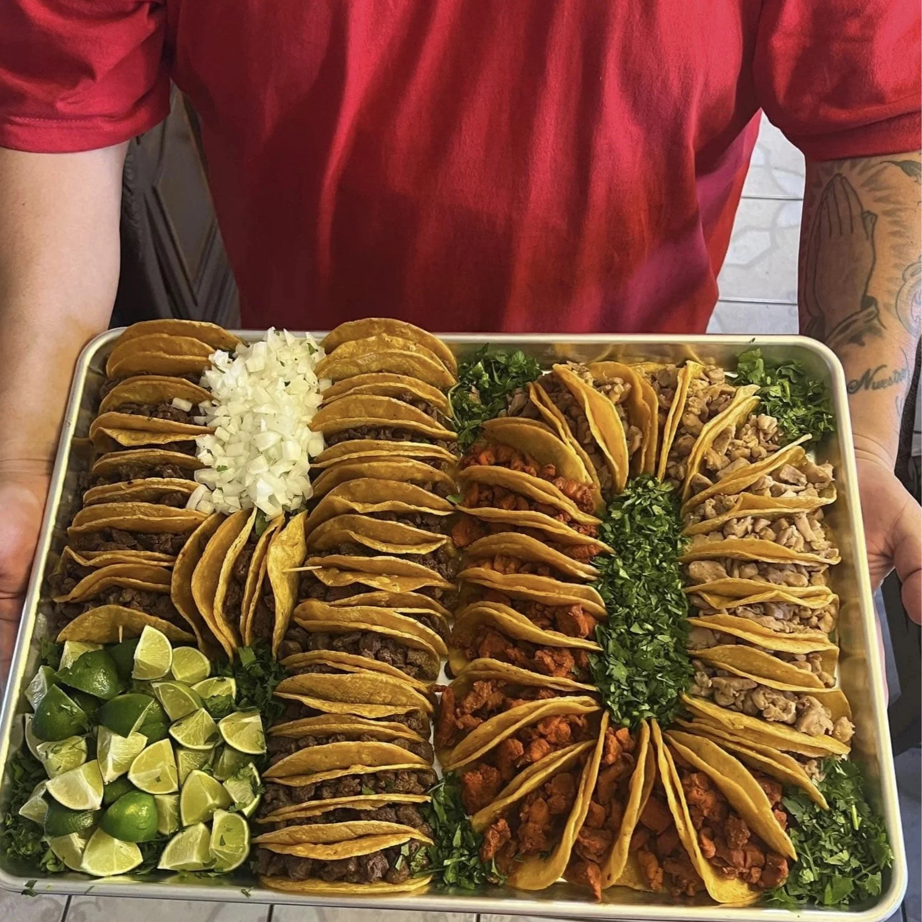 Tray of assorted tacos topped with chopped onions, lime wedges, and cilantro held by a person in a red shirt.