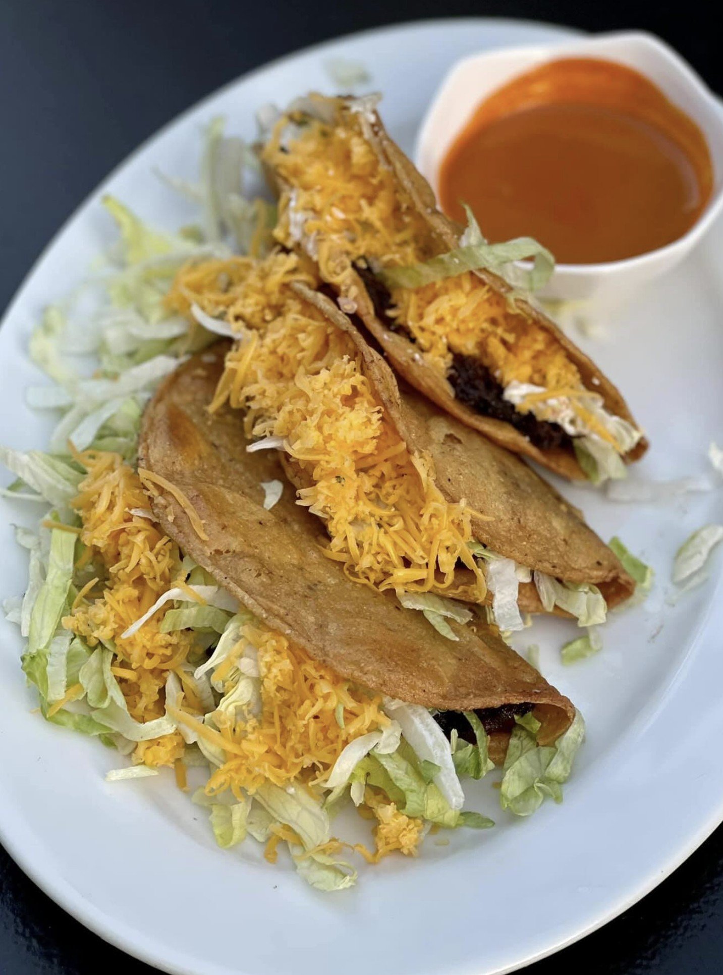 Three tacos filled with lettuce, shredded cheese, and possibly beef, on a white plate, with a side of red sauce.