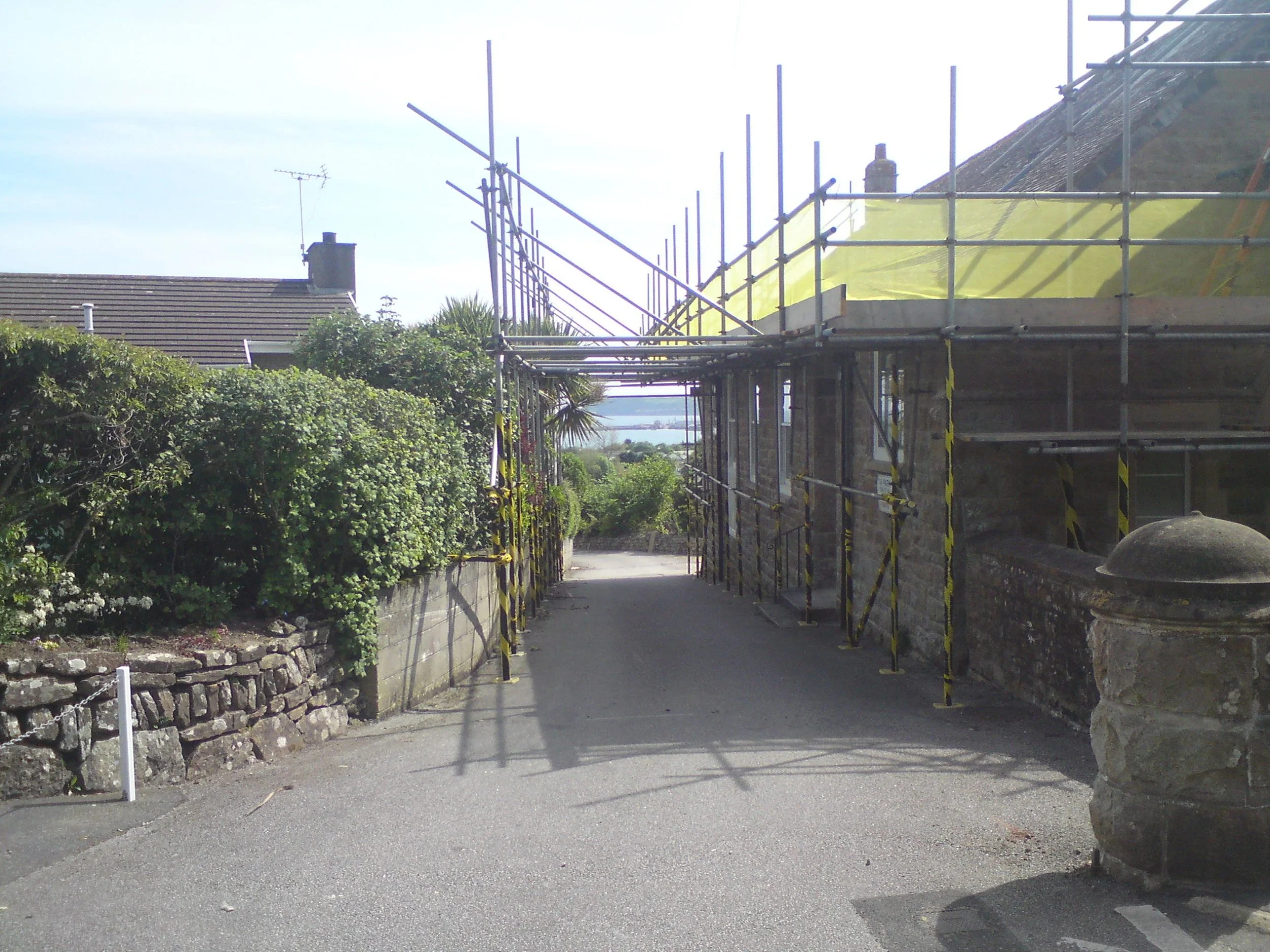 Narrow alley with ongoing construction, surrounded by greenery and stone walls, with scaffolding and safety netting.