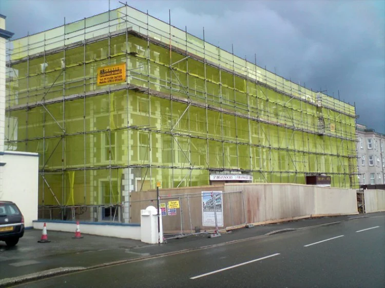 Construction site with a building covered in yellow construction wrap, surrounded by scaffolding and a wooden barrier, on a street with a parked car and traffic cones.