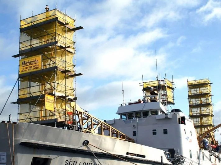 A large ship named 'Scillonian' docked near a construction site with four scaffolding towers covered in yellow safety netting, set against a partly cloudy sky.