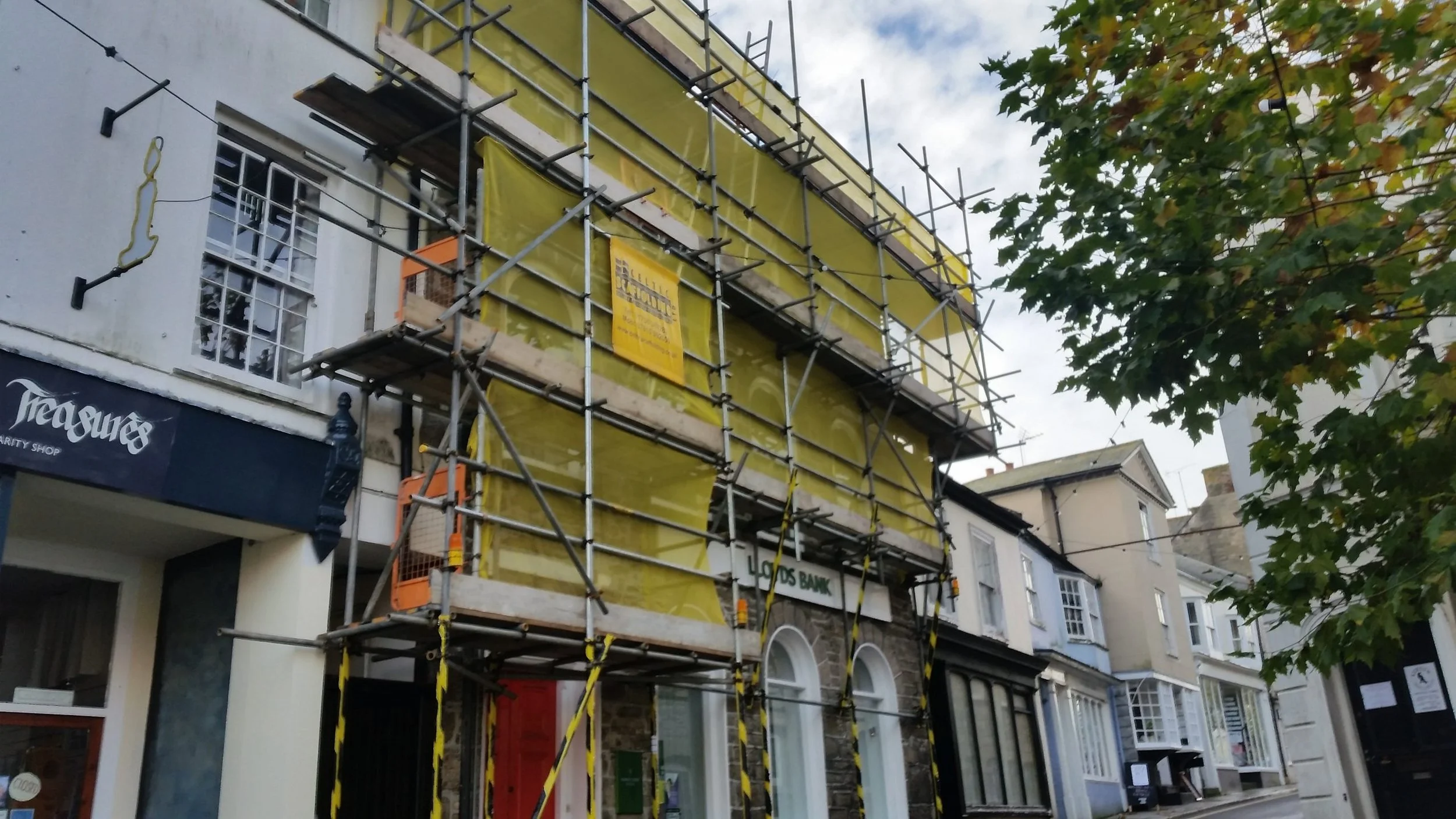 A building undergoing construction with yellow plastic sheeting covering it, surrounded by metal scaffolding. Adjacent shops with awnings are visible on the street, and there are trees with green leaves on the right side.
