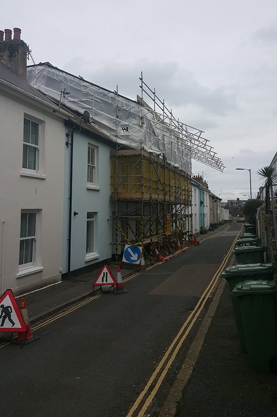 A street with houses under construction, scaffolding up against the building with protective white plastic sheeting. Traffic cones and signs indicate construction work, and trash bins are lined up along the street.