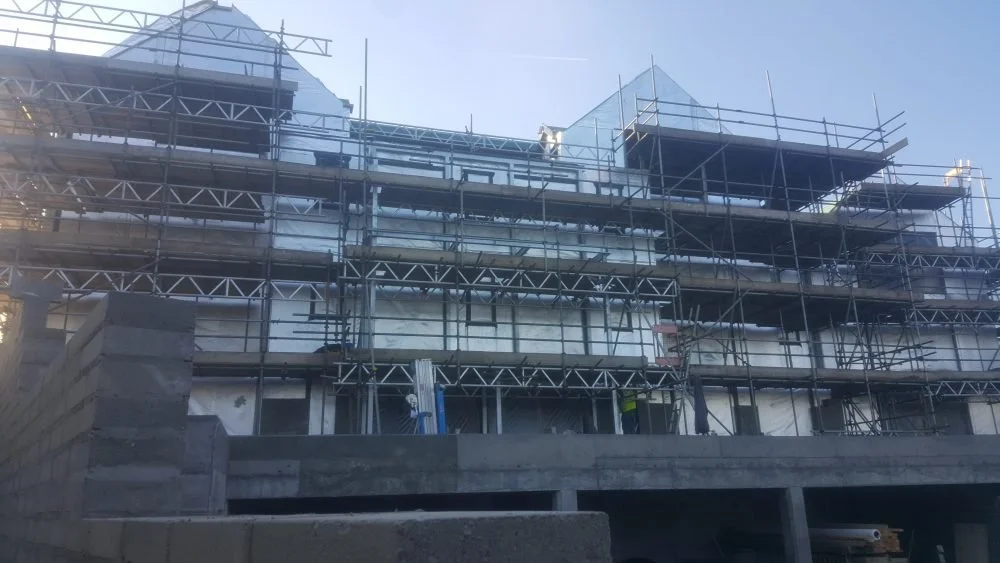 Construction workers on scaffolding surrounding a multi-story building under construction, with a concrete foundation and partly finished walls.