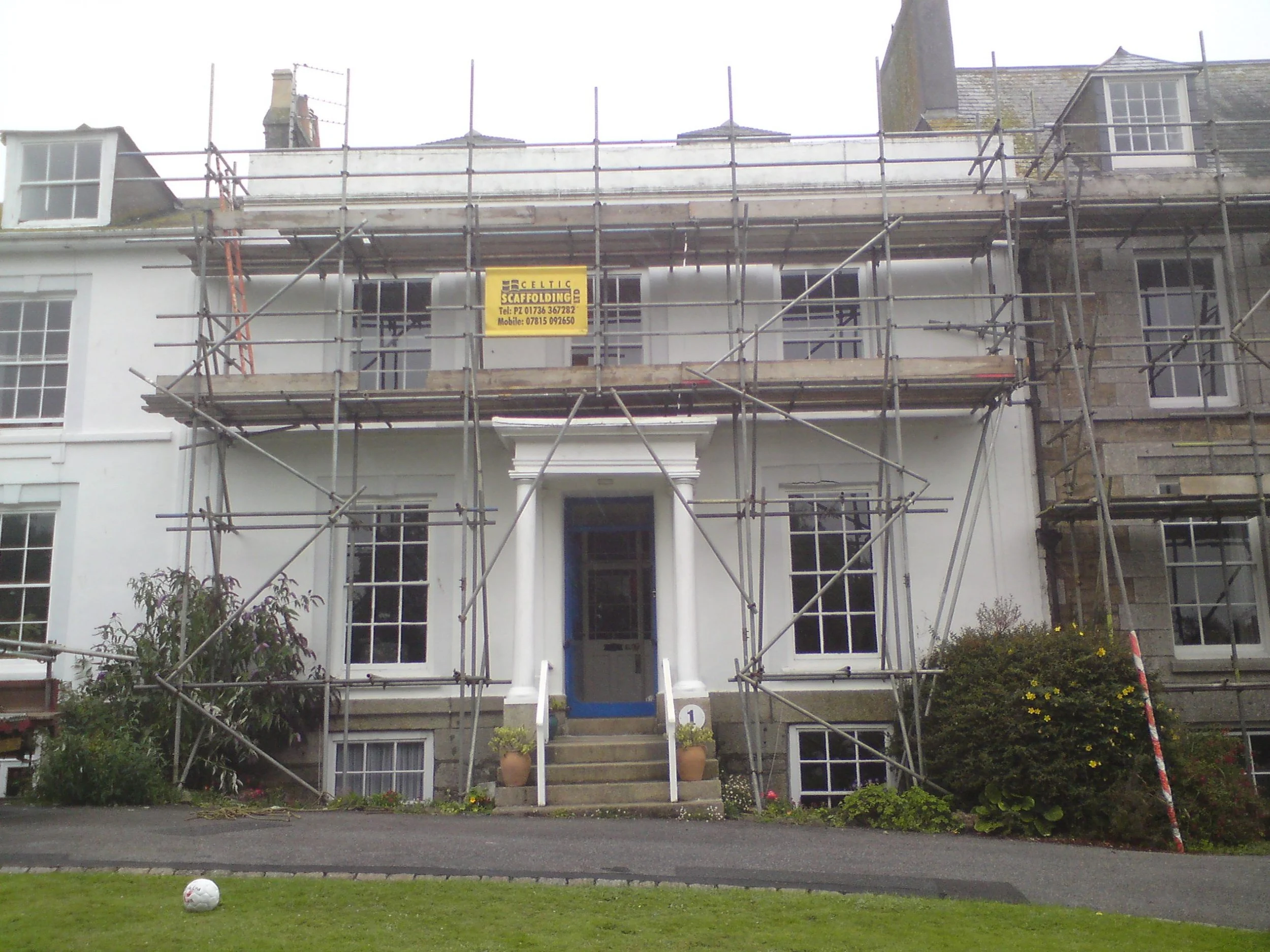 House under renovation with scaffolding around it, white facade, front steps, and gardening outside.