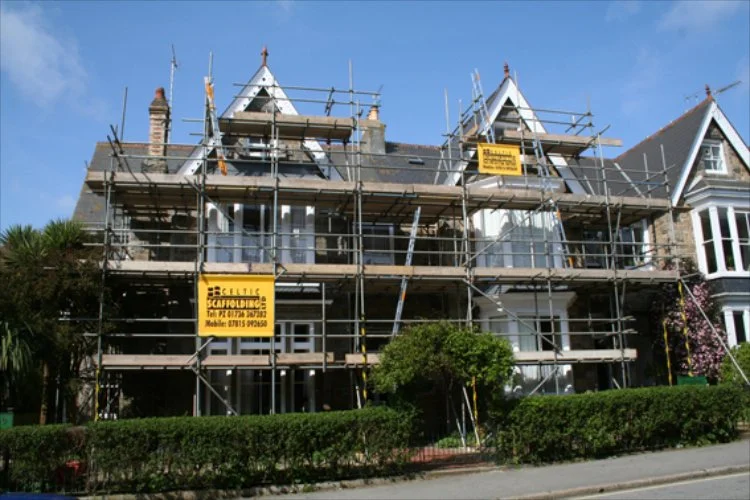 A large house undergoing renovation with scaffolding all around it, and signs indicating construction work.