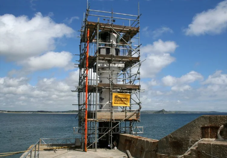 A lighthouse surrounded by scaffolding and safety signs, situated on a concrete pier by the ocean with cloudy sky overhead.