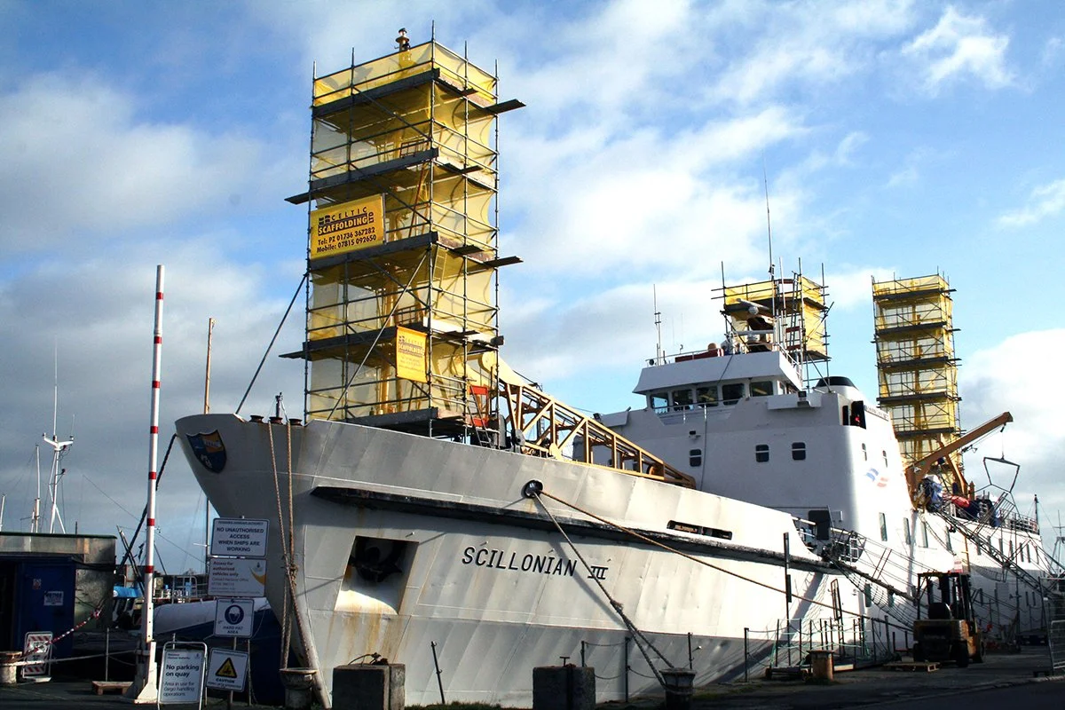 A large ship called SCILLONIAN IV is docked at a port under a partly cloudy sky. The ship is undergoing maintenance or construction with scaffolding wrapped around parts of the vessel.