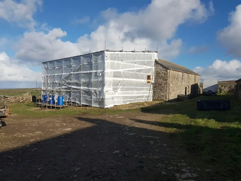 A rural scene with an old stone building partially covered in construction scaffolding and white protective sheeting, under a blue sky with clouds.