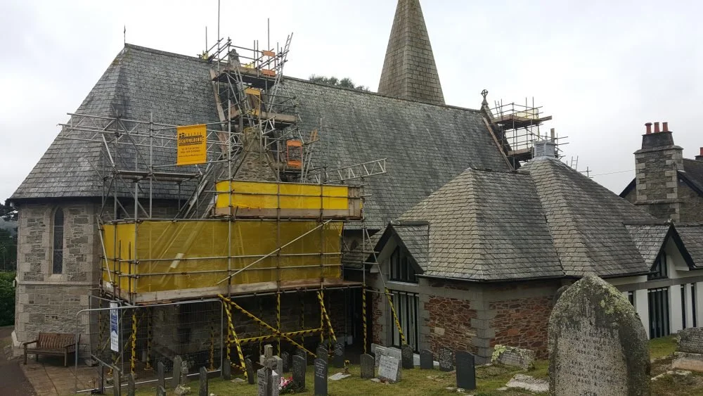 A church surrounded by scaffolding for repairs, with a graveyard in the foreground.