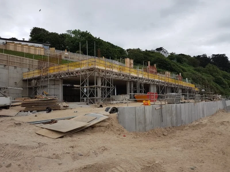 Construction site with scaffolding and concrete structures near a hillside with trees under cloudy sky.
