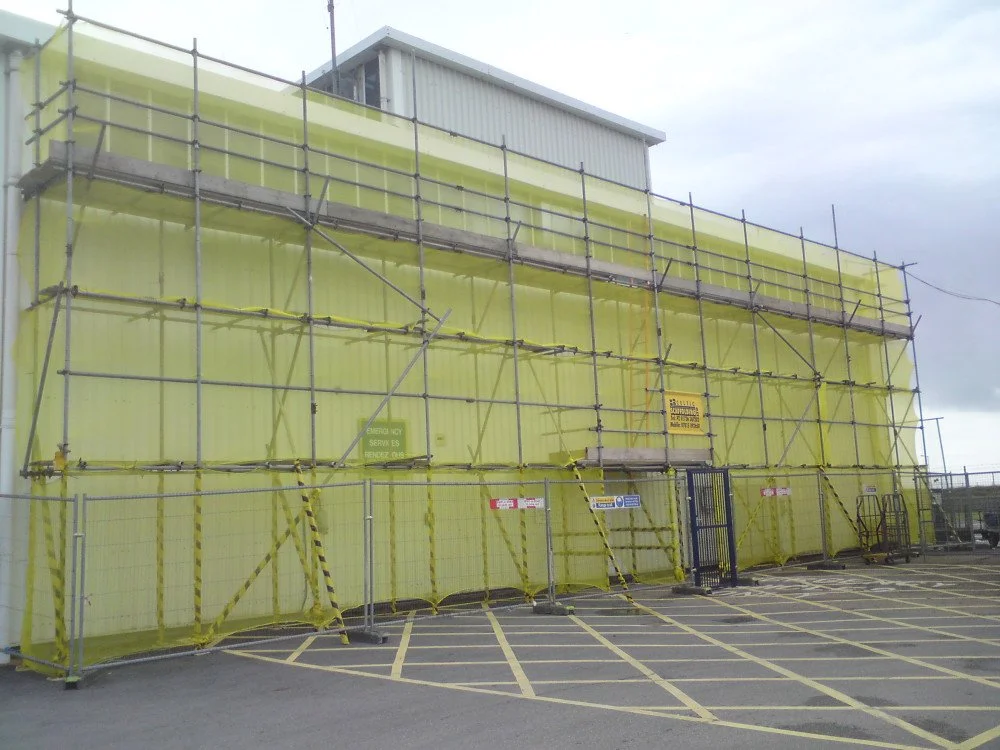 Building under construction wrapped in yellow safety netting with metal scaffolding and construction signs, in a parking lot.