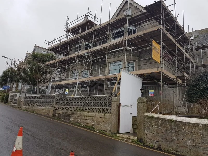A large, multi-story building under construction, surrounded by scaffolding. The building has a stone exterior, and there is a white door with a small fence and a stone wall in front. Construction signs are posted, and orange cones are on the street.