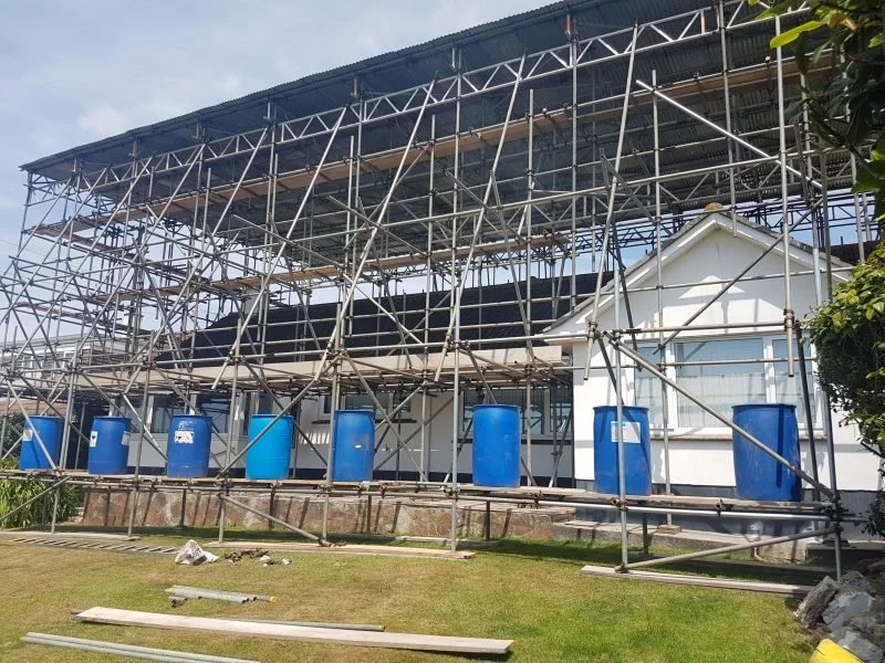 Scaffolding erected around a white house under construction or renovation, with blue barrels aligned at the base, on a grassy yard, under a partly cloudy sky.