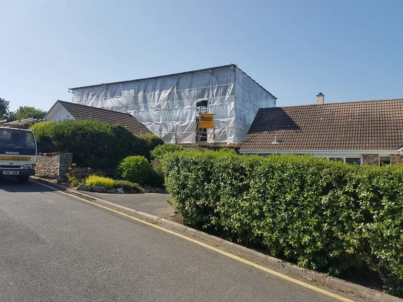 A house under renovation covered with scaffolding and white tarp, with a renovation sign on the scaffolding, next to a hedge and parked vehicle on a sloped street.